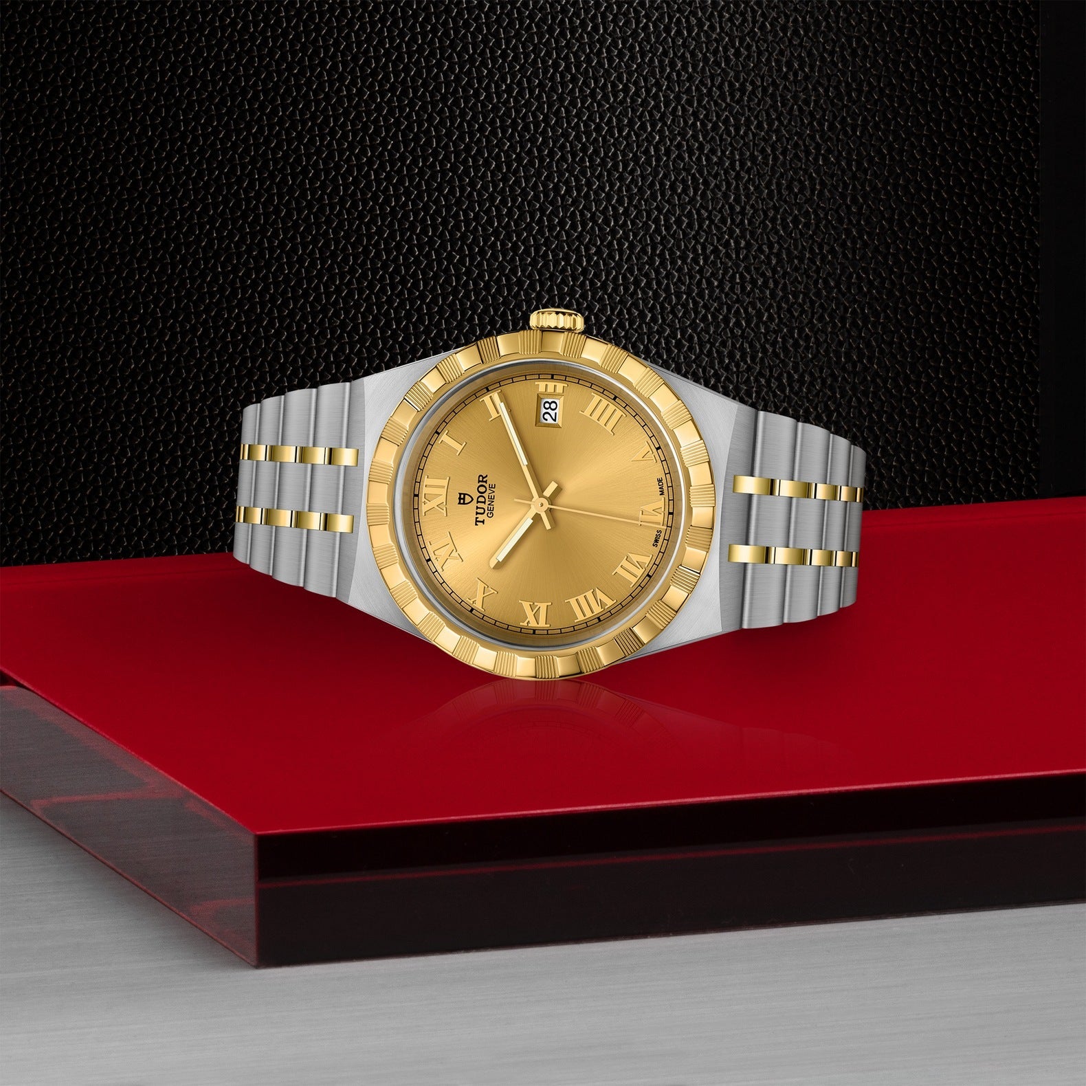 A TUDOR Royal watch by TUDOR, crafted in stainless steel and gold with a Roman numeral face and date display, rests elegantly on a red surface against a textured black backdrop.