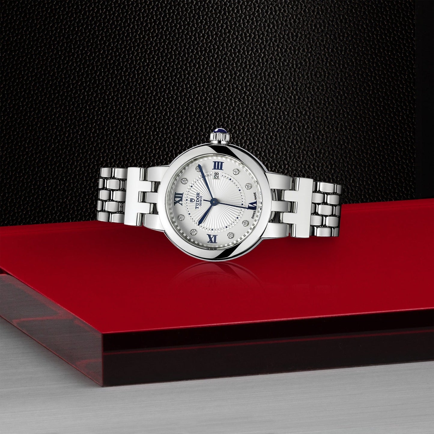 The TUDOR Clair de Rose, a luxury silver watch with a round face showcasing Roman numerals and diamond markers, is elegantly displayed on a glossy red surface. Its polished steel bracelet radiates sophistication, complemented by a small blue gemstone that crowns its design. A textured black background further elevates the watch's timeless allure.