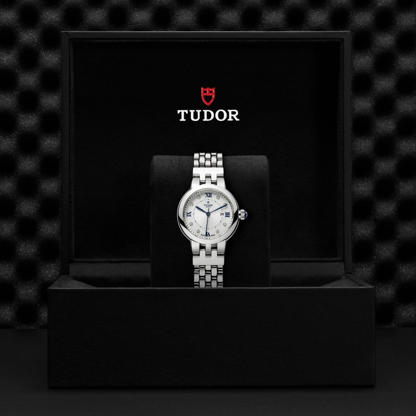 A silver TUDOR Clair de Rose wristwatch with a white opaline dial is shown in an open black box that displays "TUDOR" and a red shield logo on the inside. The watch, which features a steel case and metallic band, is set against a textured black background.