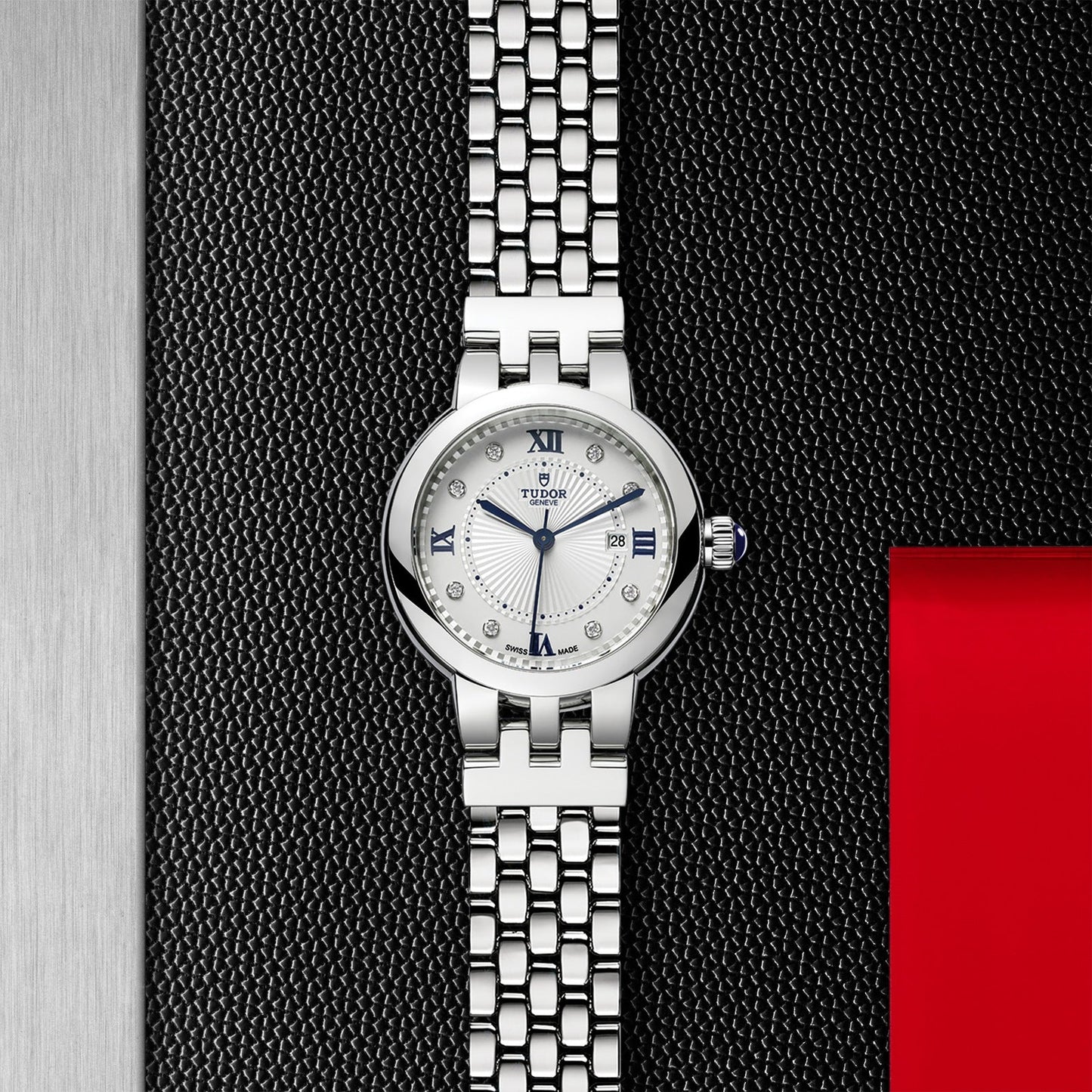 The TUDOR Clair de Rose by TUDOR is a silver wristwatch featuring a white opaline dial with blue lacquered Roman numerals and a date display at 3 o'clock. It boasts an elegant metal link band set against a textured dark background, accentuated by a subtle red rectangular detail.