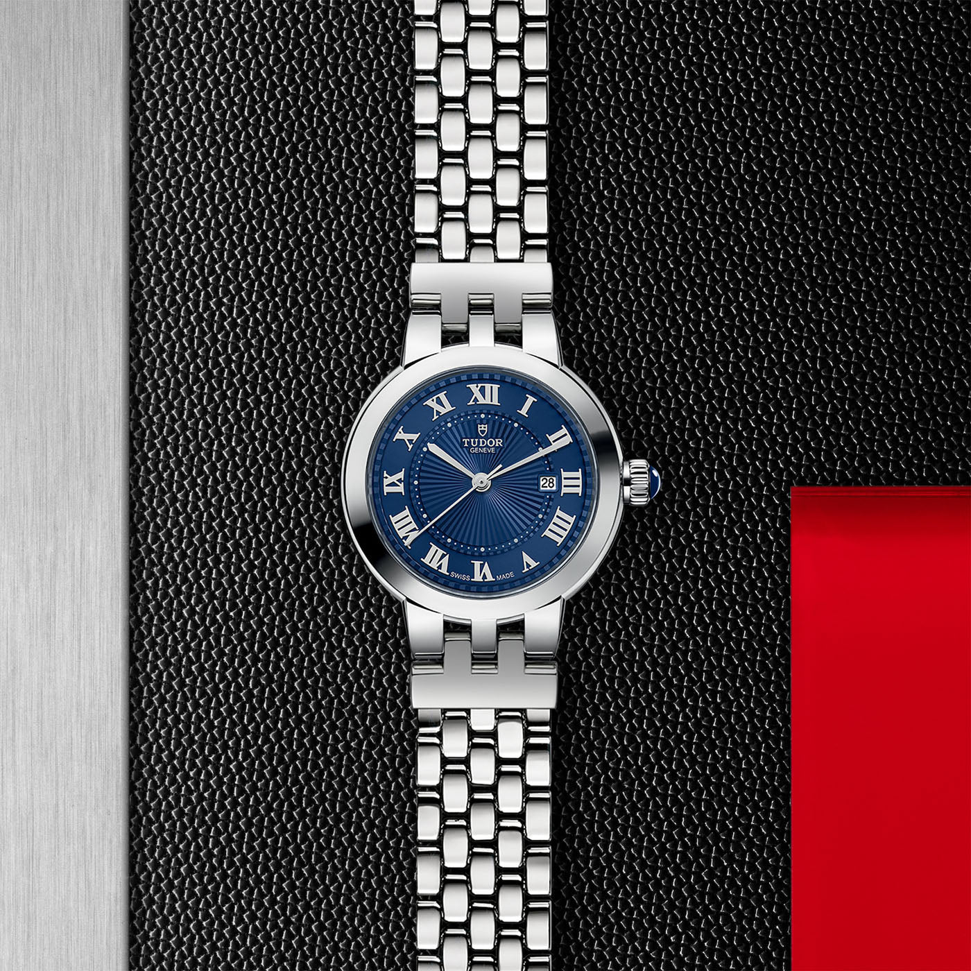 The TUDOR Clair de Rose wristwatch, featuring a blue face with Roman numerals and a stylish steel bracelet, is elegantly showcased on a textured black surface accented with red and silver tones.