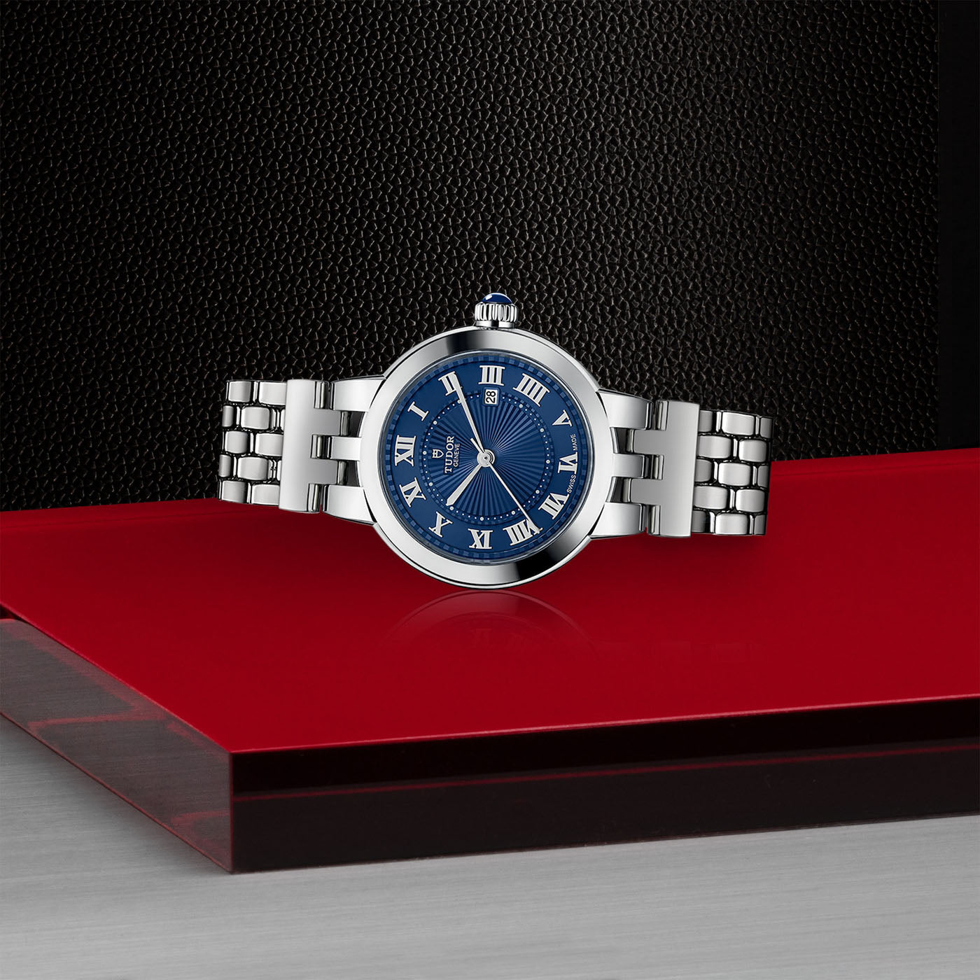 The TUDOR Clair de Rose wristwatch, featuring a silver design with an opaline blue face and Roman numerals, is elegantly showcased against a striking red and black background. It boasts a robust steel bracelet and a round case, combining sophistication with timeless style.