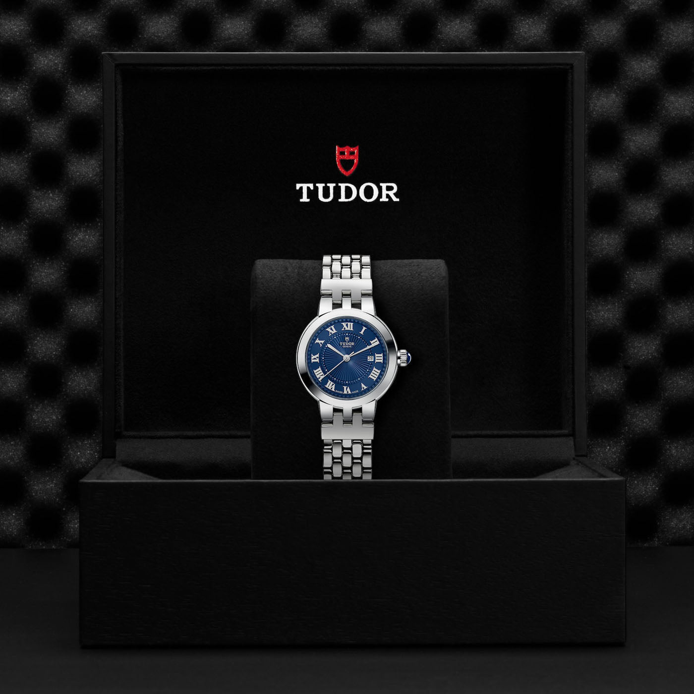 The TUDOR Clair de Rose, a luxury watch crafted by TUDOR, showcases a blue dial with Roman numerals and elegant hands. It comes with a steel bracelet and is presented in an exquisite black box. The timeless sophistication of the watch is further highlighted by the visible TUDOR logo above it inside the box.