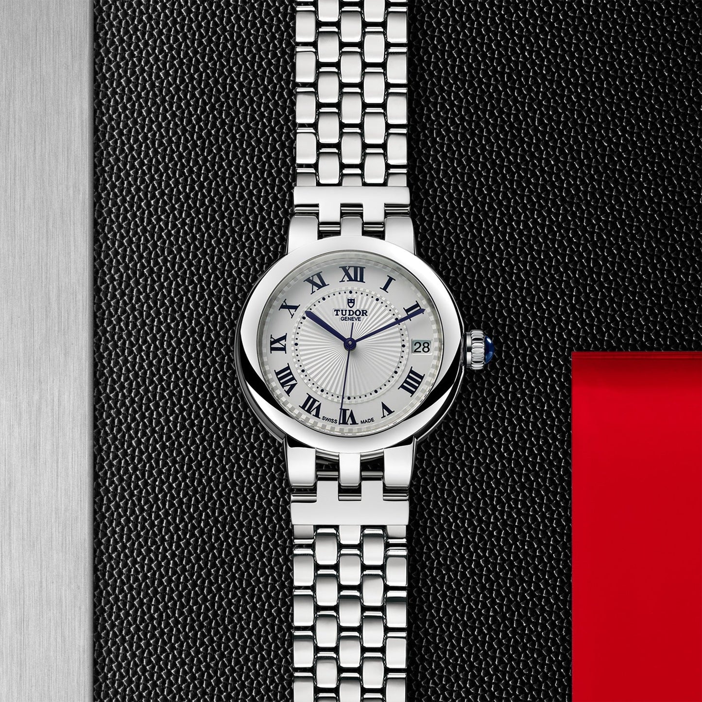 The TUDOR Clair de Rose watch, featuring a sleek silver design and a woven metal band, is elegantly showcased against a textured black and red backdrop. Its steel case houses an opaline dial embellished with Roman numerals and a date indicator, radiating timeless sophistication.