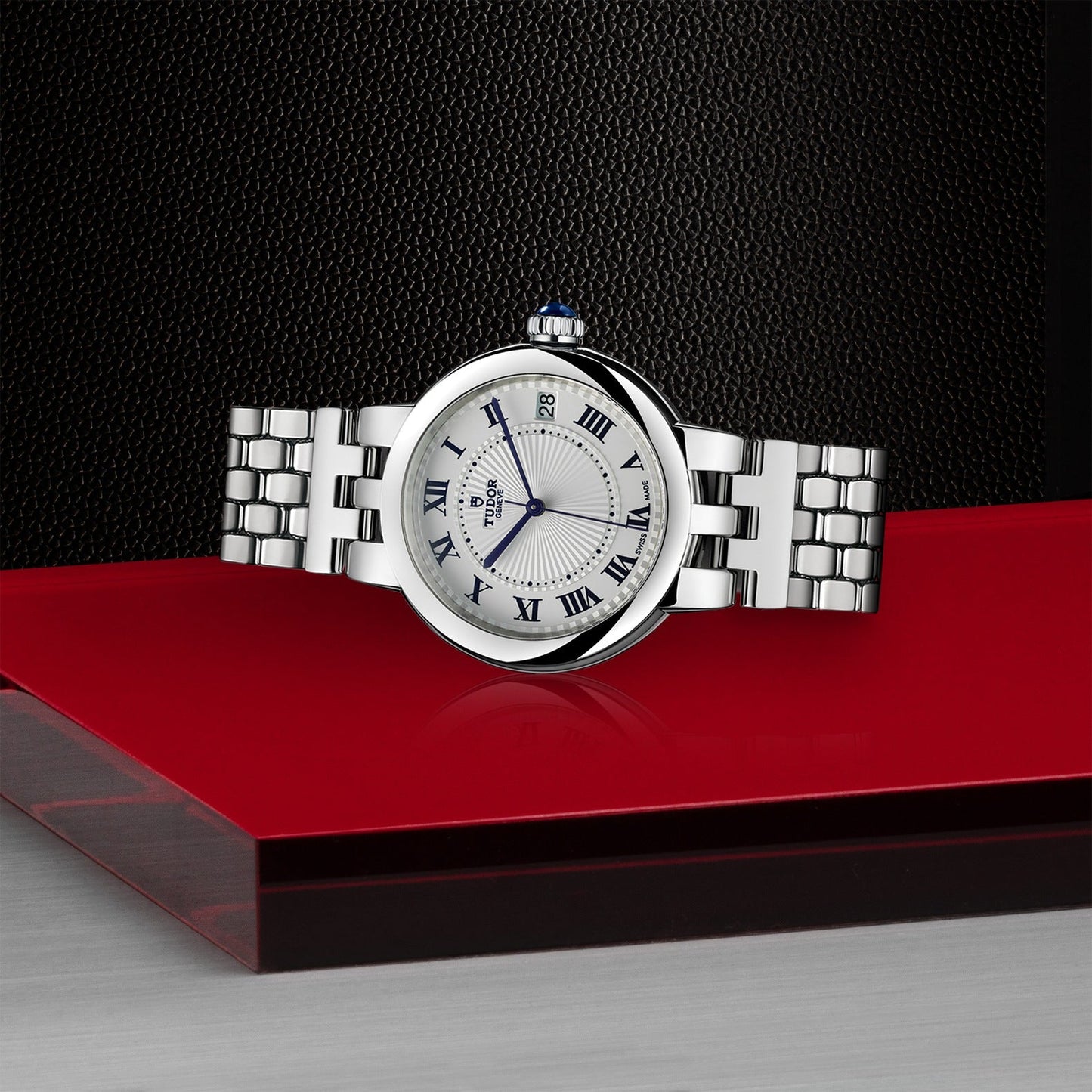 A TUDOR Clair de Rose wristwatch, featuring a polished metal bracelet, a blue-tipped crown, and an elegant steel case, with a white opaline dial and Roman numerals, is placed on a red and black geometric surface.