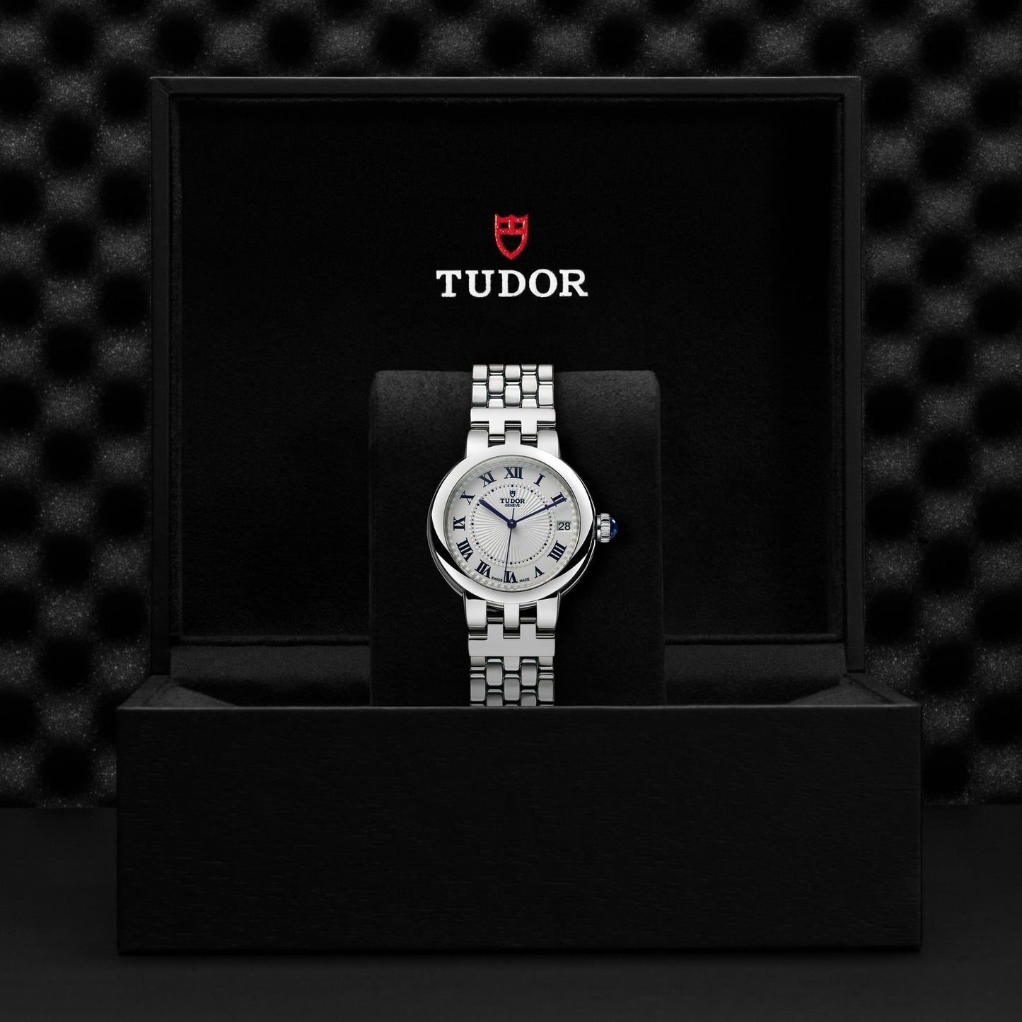 A TUDOR Clair de Rose wristwatch featuring a steel case, silver band, and opaline dial with Roman numerals is showcased in a black velvet-lined box. The TUDOR logo is elegantly displayed on the inner lid above the watch.