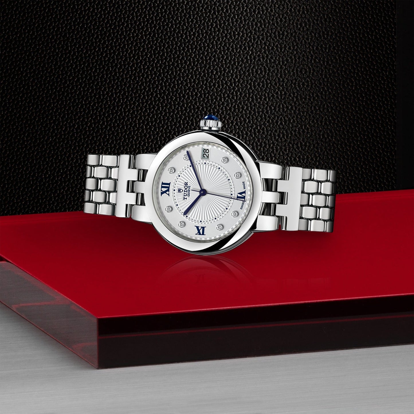 A TUDOR Clair de Rose luxury wristwatch with an opaline dial and a round white face is elegantly displayed on a glossy red surface. It features Roman numerals, sapphire crown, and a steel bracelet, set against a textured black background.