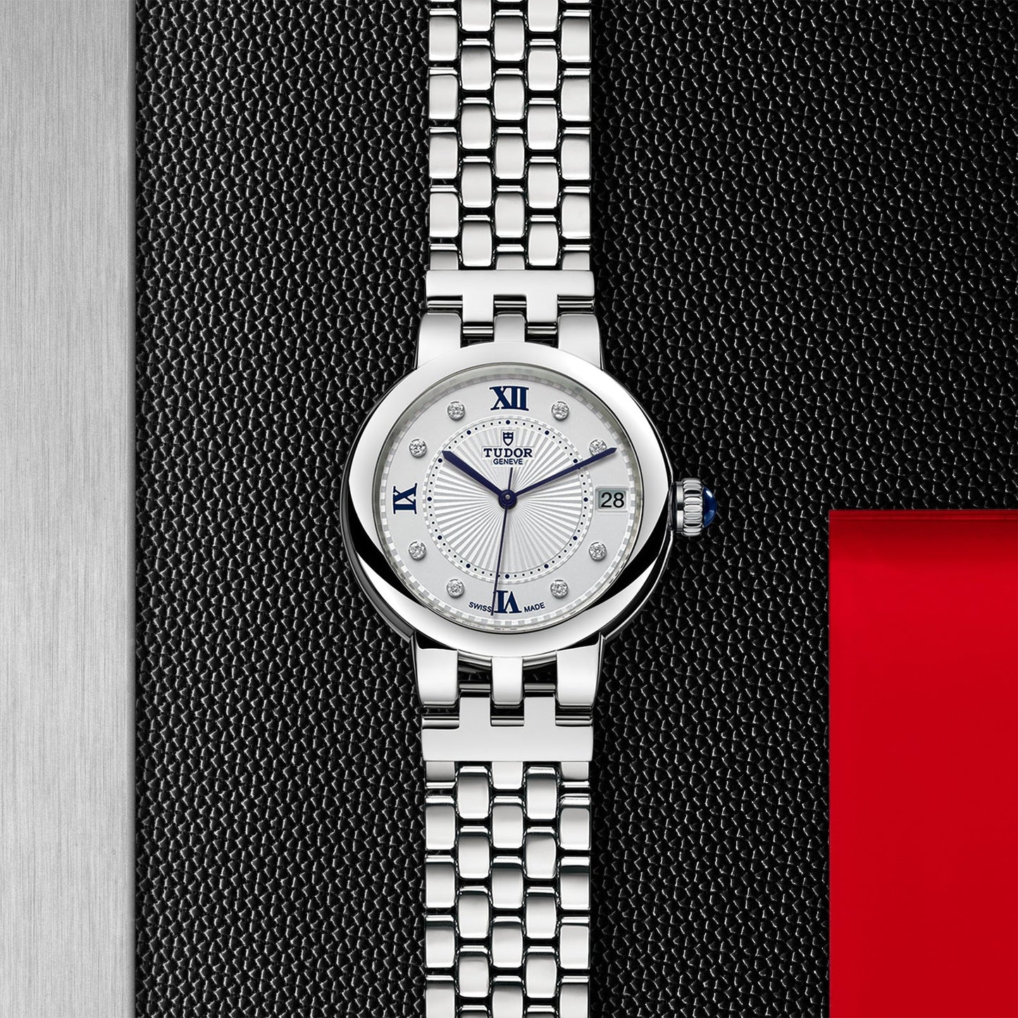 The TUDOR Clair de Rose wristwatch features a silver finish with a patterned metal band and an opaline dial. It elegantly displays Roman numerals and diamond hour markers, set against a backdrop that includes a black textured surface, a red square, and a metallic vertical strip, embodying the sophisticated TUDOR style.