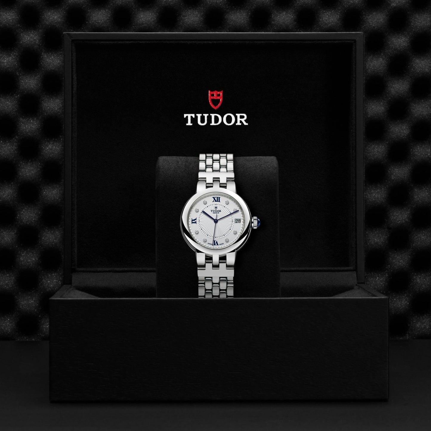 The TUDOR Clair de Rose, a silver wristwatch with a round face displaying Roman numerals, is nestled in a black box. The red and white TUDOR logo is positioned above the watch. Its opaline dial adds an elegant touch against the textured dark background.