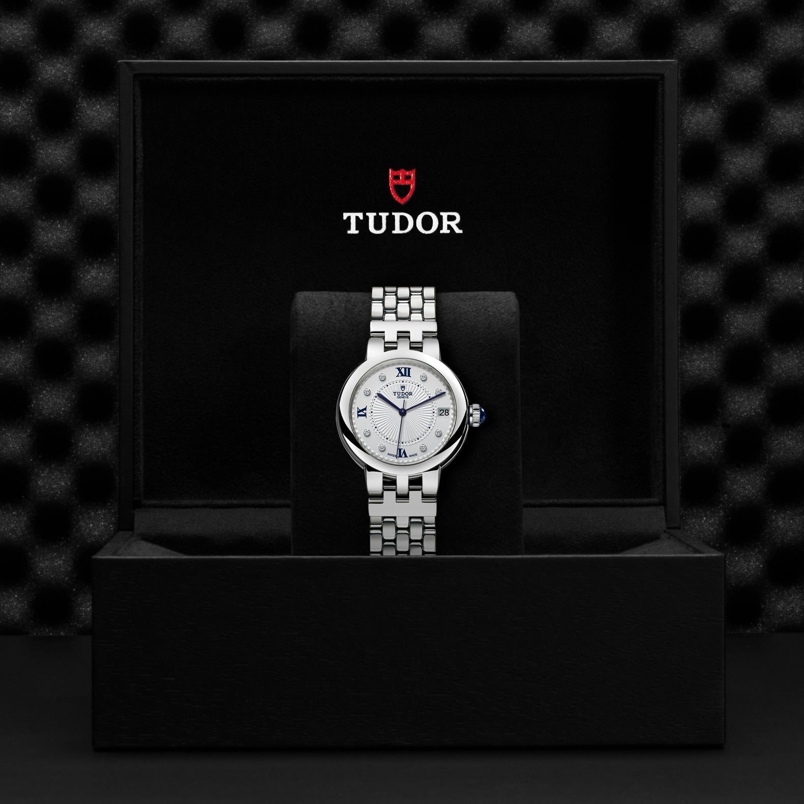 The TUDOR Clair de Rose, a silver wristwatch with a round face displaying Roman numerals, is nestled in a black box. The red and white TUDOR logo is positioned above the watch. Its opaline dial adds an elegant touch against the textured dark background.