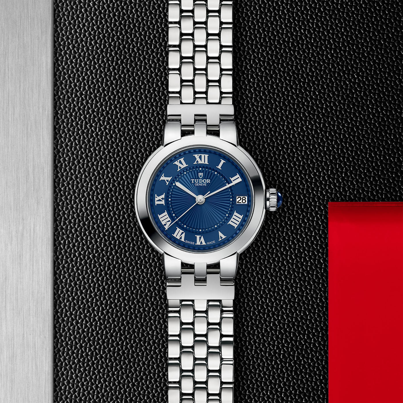 Displayed on a textured dark surface is the TUDOR Clair de Rose, a silver wristwatch with a blue face featuring Roman numerals and opaline dials. The steel bracelet adds elegance, while a small date window at the 3 o'clock position completes this timeless piece from TUDOR.