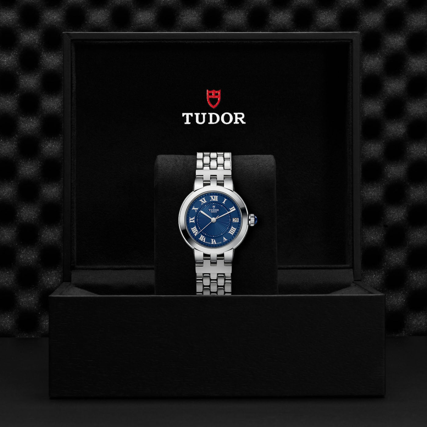 The TUDOR Clair de Rose, a silver wristwatch featuring a blue dial and Roman numerals, is elegantly showcased with its sleek steel bracelet in an open black box adorned with the "TUDOR" inscription inside. The ensemble is set against a textured black background.