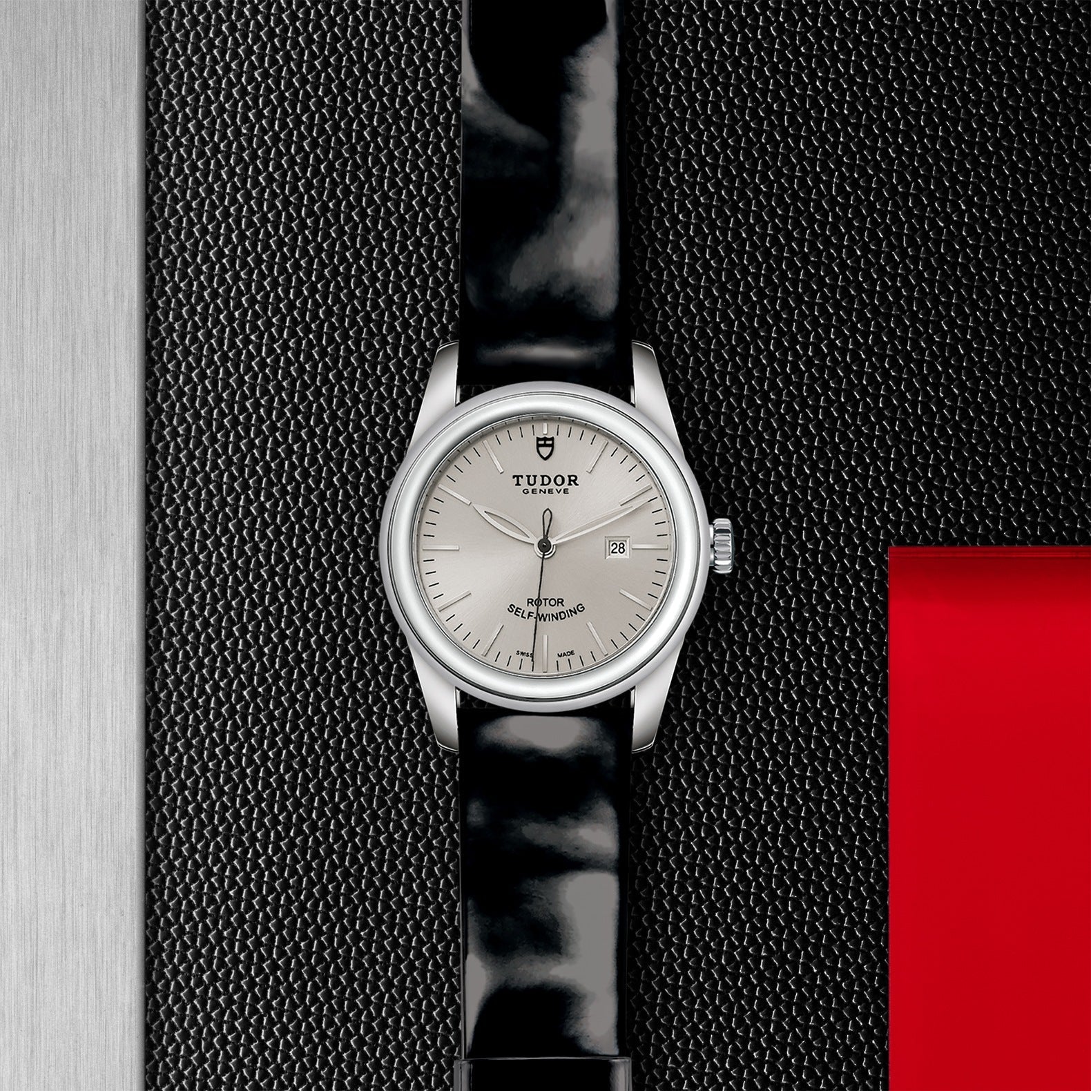 Displayed against a textured dark background with a striking red accent on the right, the New Old Stock TUDOR Glamour Date watch features an elegant silver face paired with a black leather strap. This timepiece exudes glamour and sophistication, prominently showcasing the TUDOR logo and a date feature.