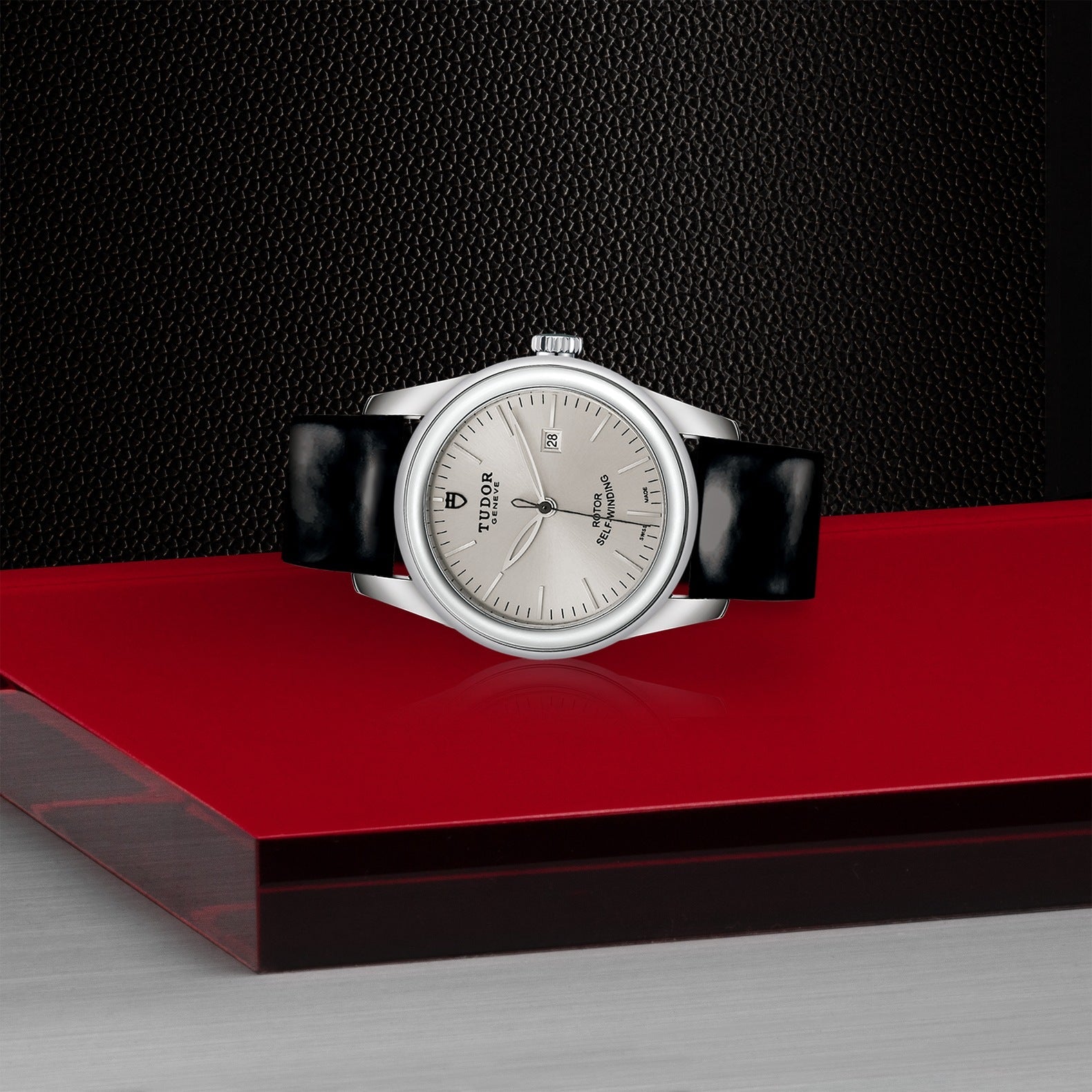 A New Old Stock TUDOR Glamour Date watch in silver, featuring a round face and black leather strap, is displayed on a glossy red platform. The background showcases a textured black surface, accentuating the watch's elegant design with an automatic Date+Day feature.