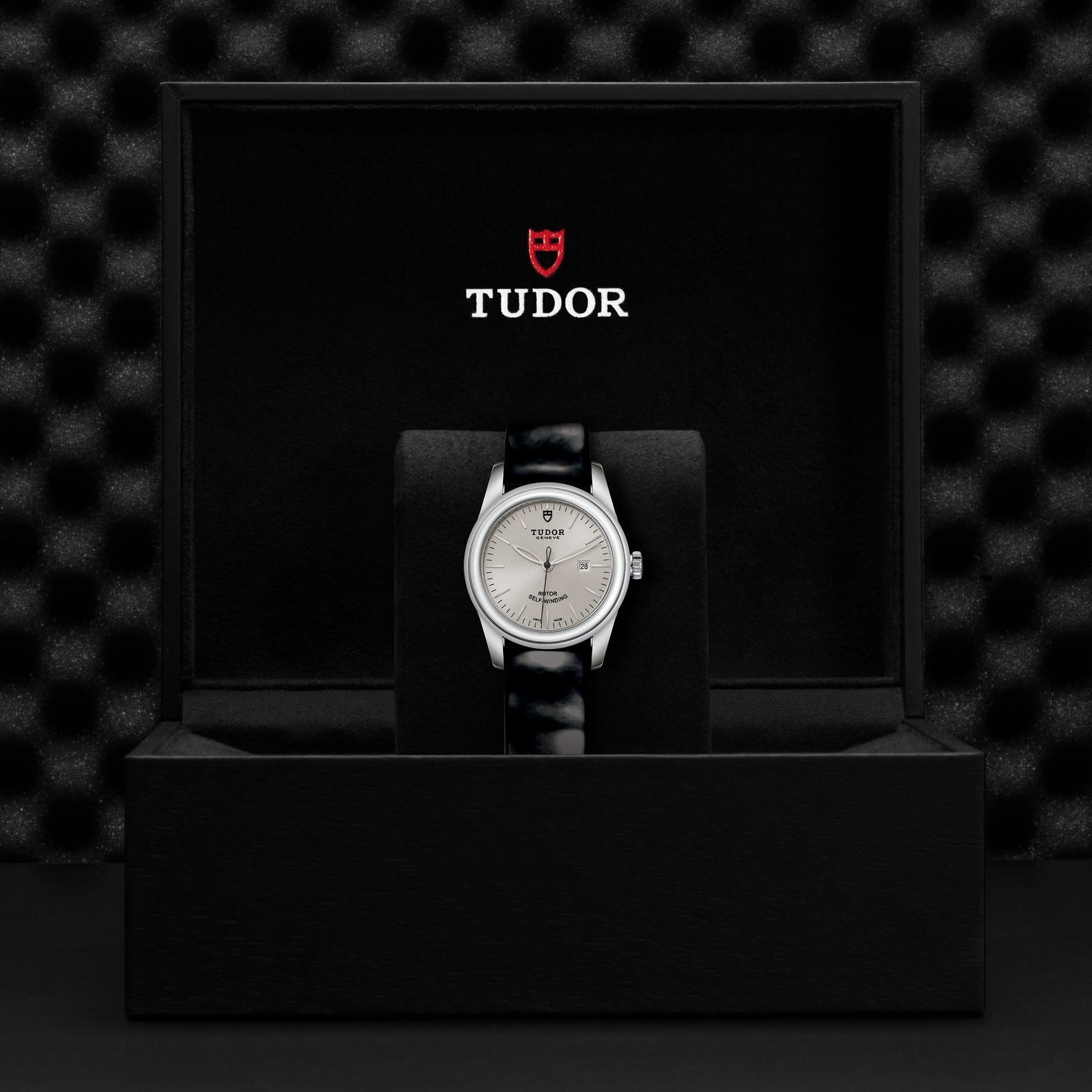 The New Old Stock TUDOR Glamour Date, featuring a silver case and a black leather strap, exudes sophistication as it is elegantly displayed in a sleek black box. The lid prominently displays the TUDOR logo in white and red, while the textured black pattern of the setting enhances the watch's glamorous appeal.