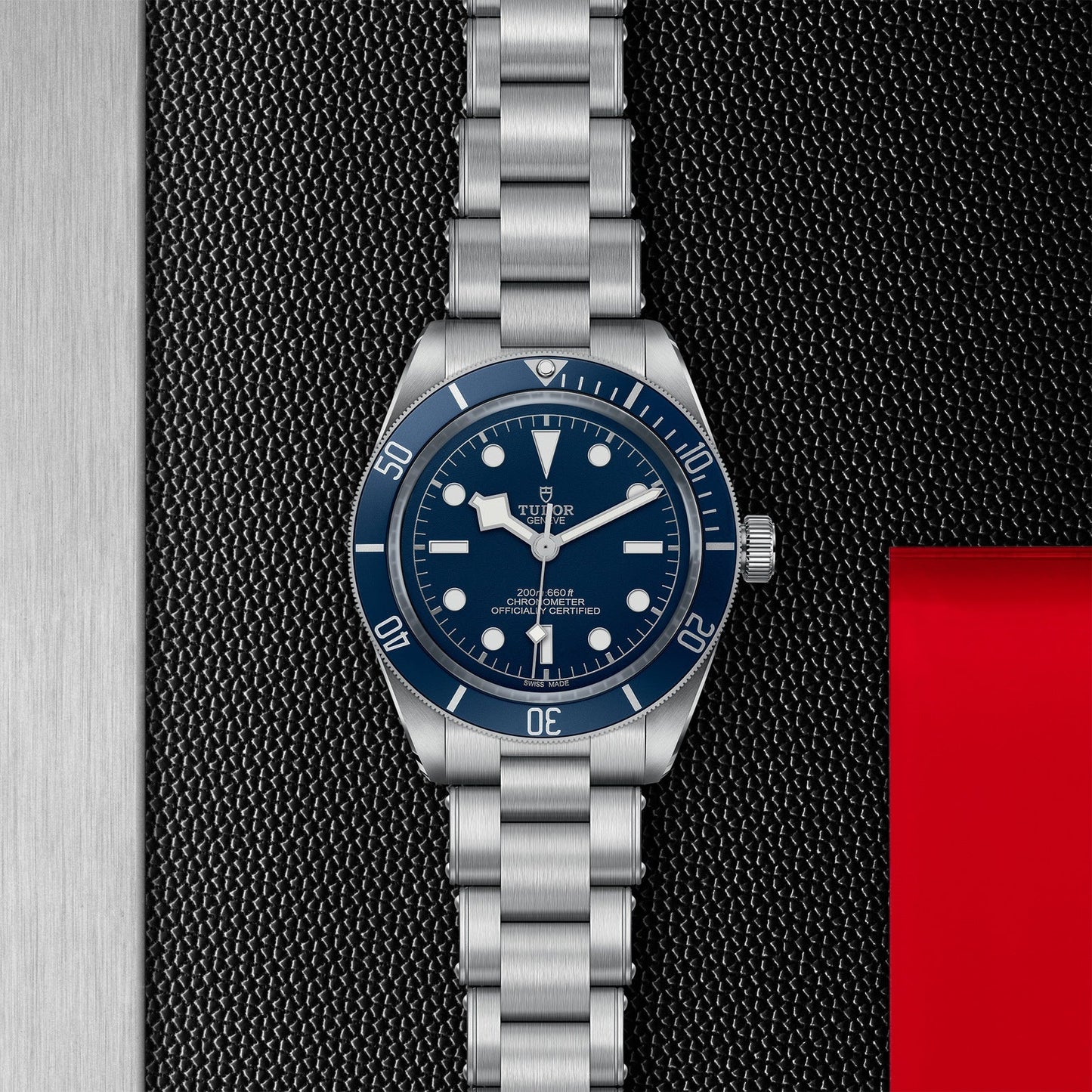 The TUDOR Black Bay 58, a dive watch with a blue dial and bezel, white hour markers, and a metal bracelet, is showcased on a textured black and gray background highlighted by a red accent on the right.