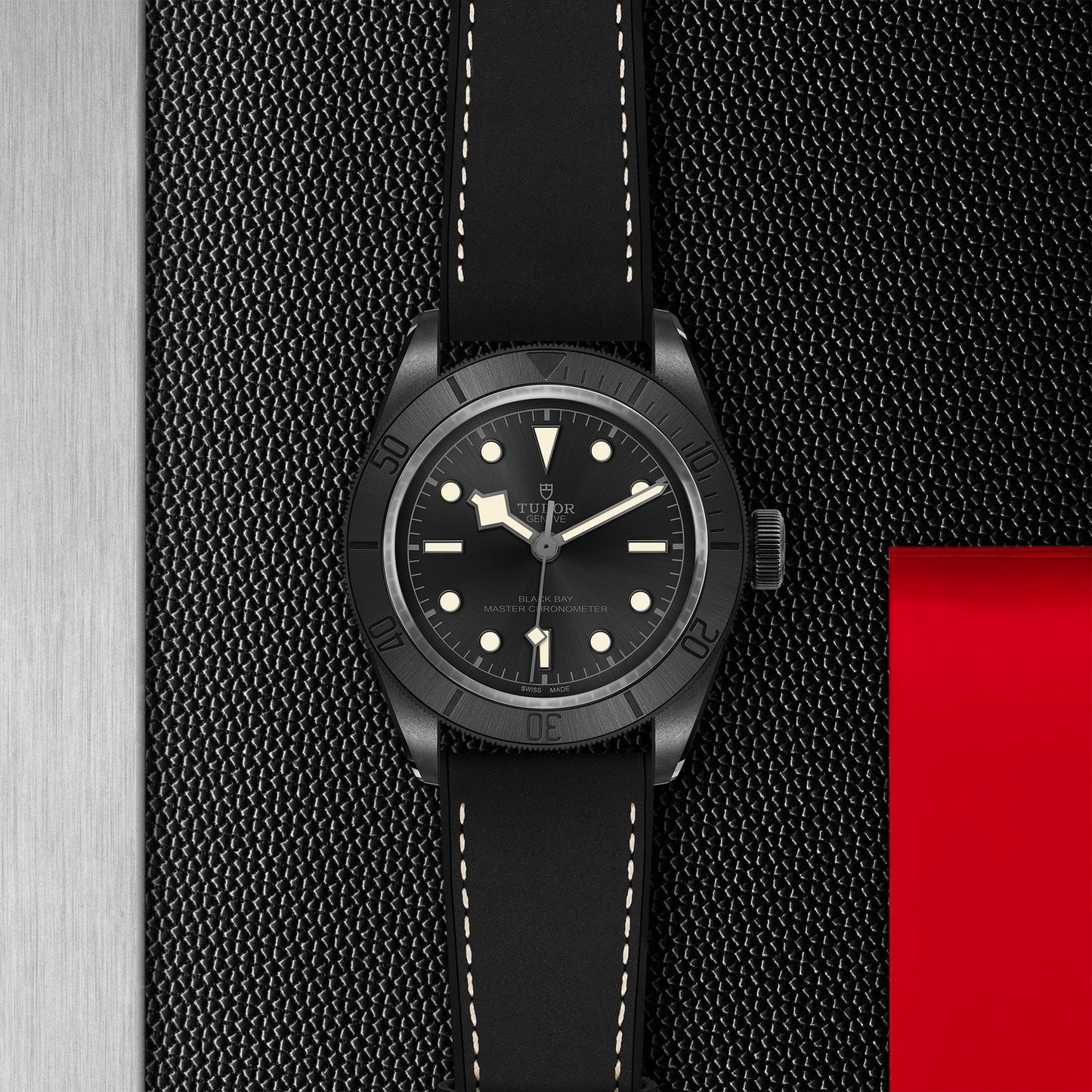 A TUDOR Black Bay Ceramic wristwatch in matte black, featuring a round dial and white hour markers, is displayed on a textured black surface. The watch includes a black strap with white stitching, complemented by vertical red and silver strips that add a modern flair to this stylish TUDOR timepiece.