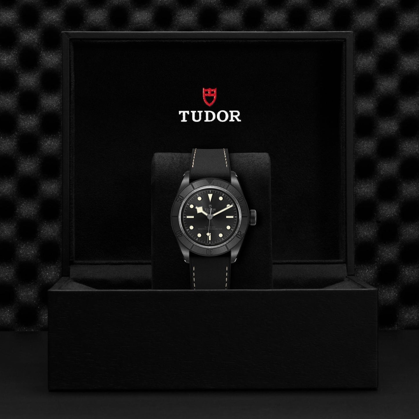 A luxury wristwatch displayed in an open black box from TUDOR. The TUDOR Black Bay Ceramic watch boasts a dark face with luminous markers, a black bezel, and a matching strap. This stunning piece offers the precision of a Master Chronometer, with the TUDOR logo elegantly showcased on the lid of the box.