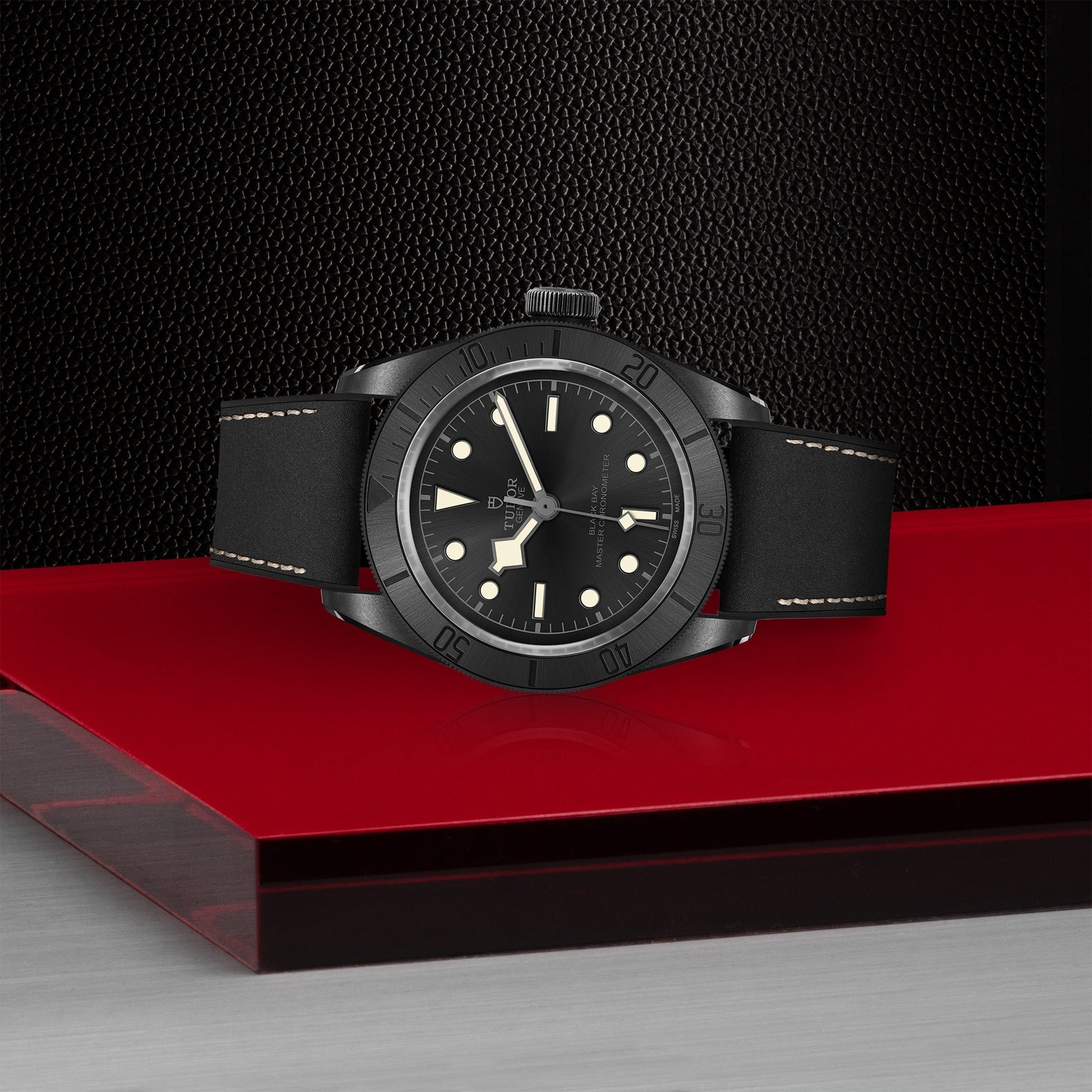 Displayed on a red and black pedestal with a textured backdrop is the TUDOR Black Bay Ceramic wristwatch. This sleek timepiece by TUDOR features a minimalistic dial with luminous hour markers, crafted from matte black ceramic. It is complemented by a black band enhanced with subtle stitching details.