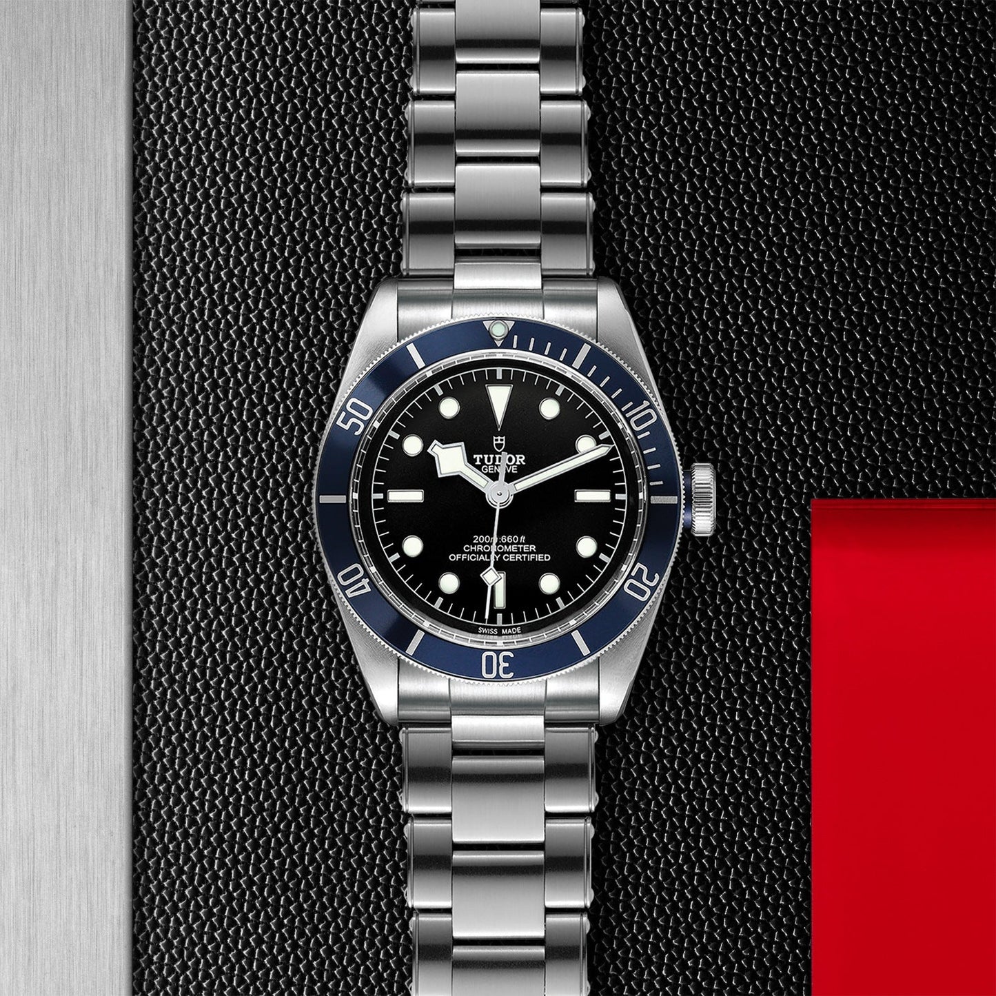 A sleek TUDOR Black Bay wristwatch with a black dial and blue bezel rests elegantly on a textured black surface. This diving watch features a stainless steel bracelet and white hour markers, complemented by a design that includes a red rectangle on the right side.