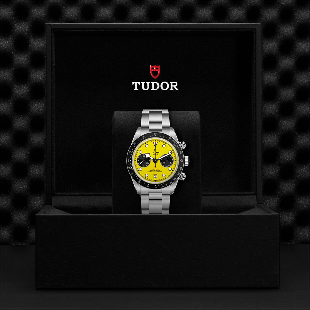 A TUDOR Black Bay Chrono 39 wristwatch with a yellow dial, black subdials, and silver 39mm case is upright in an open black TUDOR box against a textured black background.