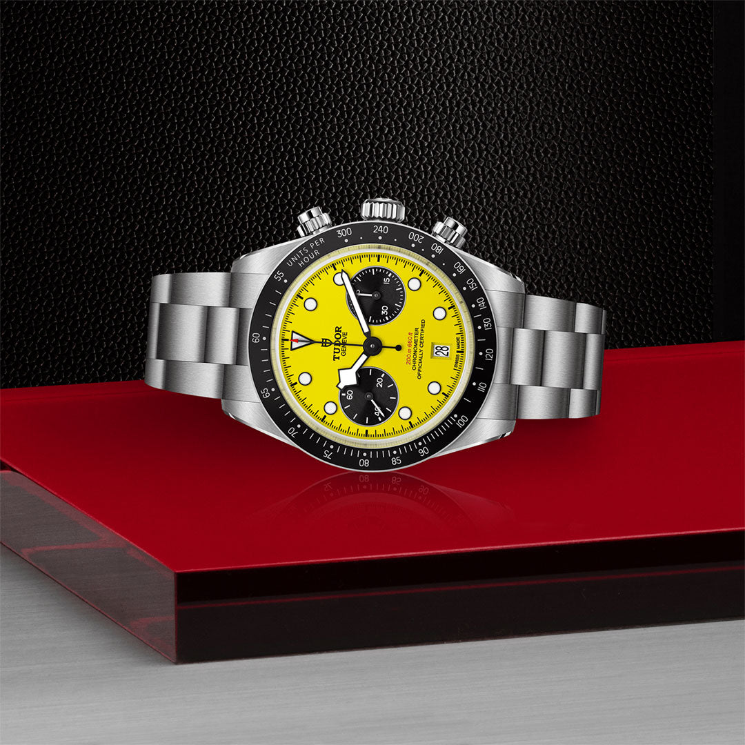 The TUDOR Black Bay Chrono 39 stainless steel chronograph watch, featuring a bright yellow dial with black subdials, is displayed on a glossy red and black platform against a textured black background.