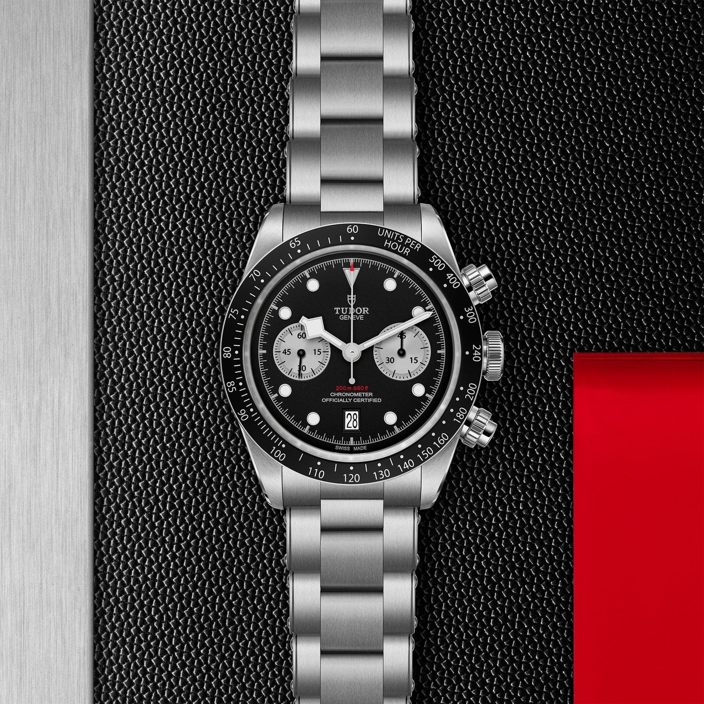 The TUDOR Black Bay Chrono by TUDOR is a sleek silver stainless steel watch, reminiscent of traditional chronographs, featuring a black dial with three sub-dials set against a textured black surface. It includes a date feature at 6 o'clock and is paired with an eye-catching red rectangular element.