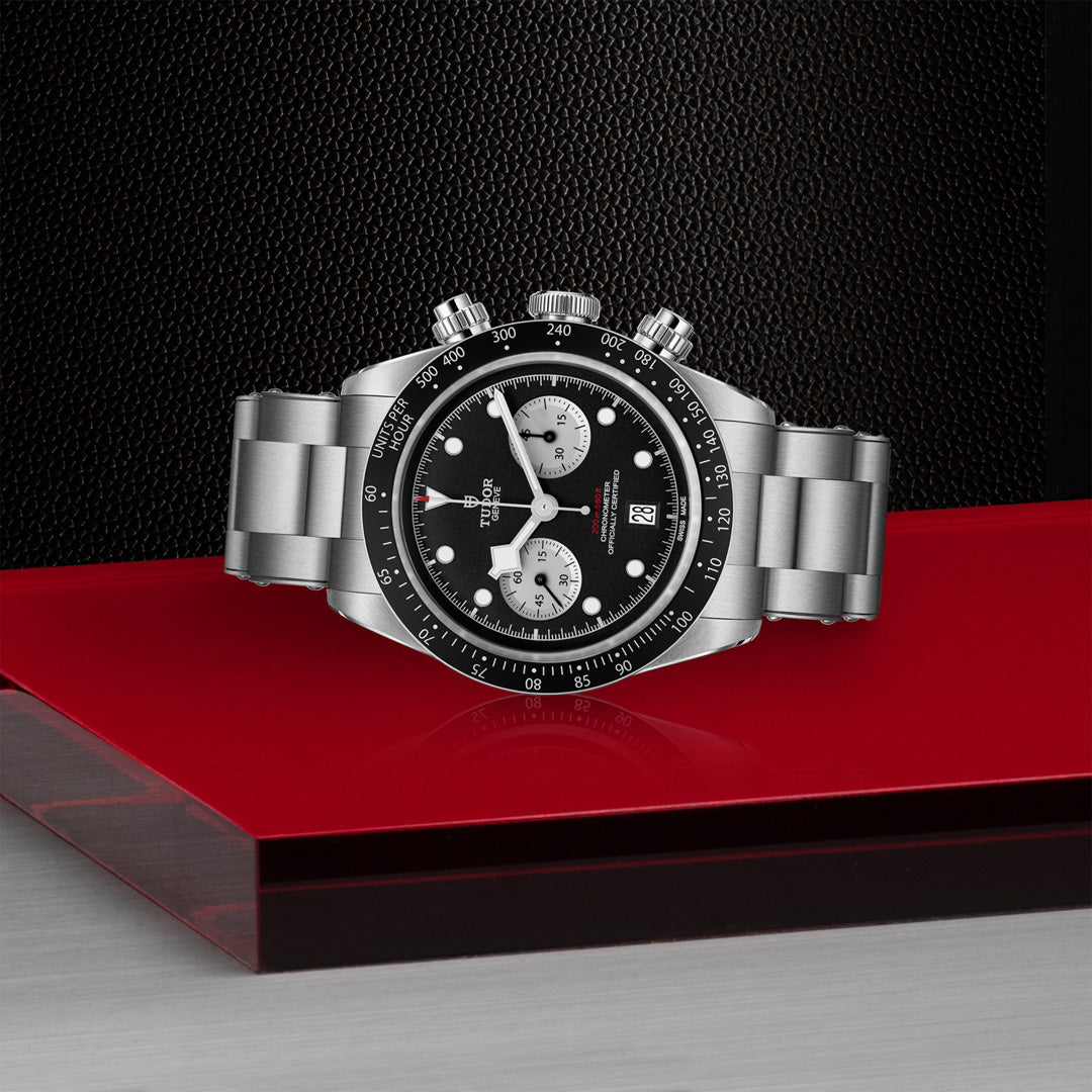 The TUDOR Black Bay Chrono, a luxury silver watch with a black face, three subdials, and a date display, is elegantly showcased on a glossy red surface with a sleek T-Fit clasp against a dark textured wall.