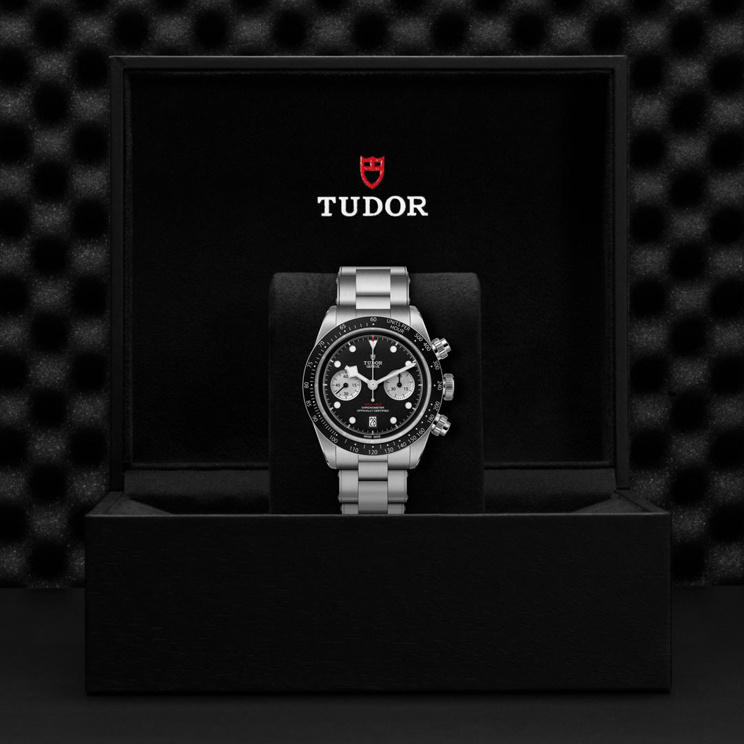 The TUDOR Black Bay Chrono, featuring a silver band and black dial, is elegantly presented in an open black box with a textured background. The stylish red and white TUDOR logo adorns the inside lid, while the T-Fit clasp ensures a precise fit.