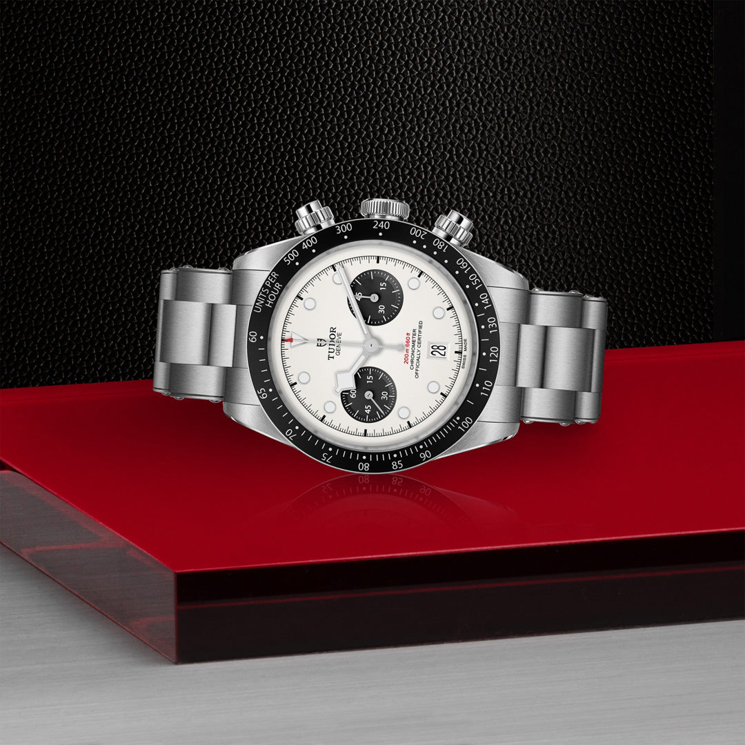A TUDOR Black Bay Chrono wristwatch with a white face, black bezel, and two black subdials rests on a glossy red surface. The background has a textured black section and a smooth silver bottom.