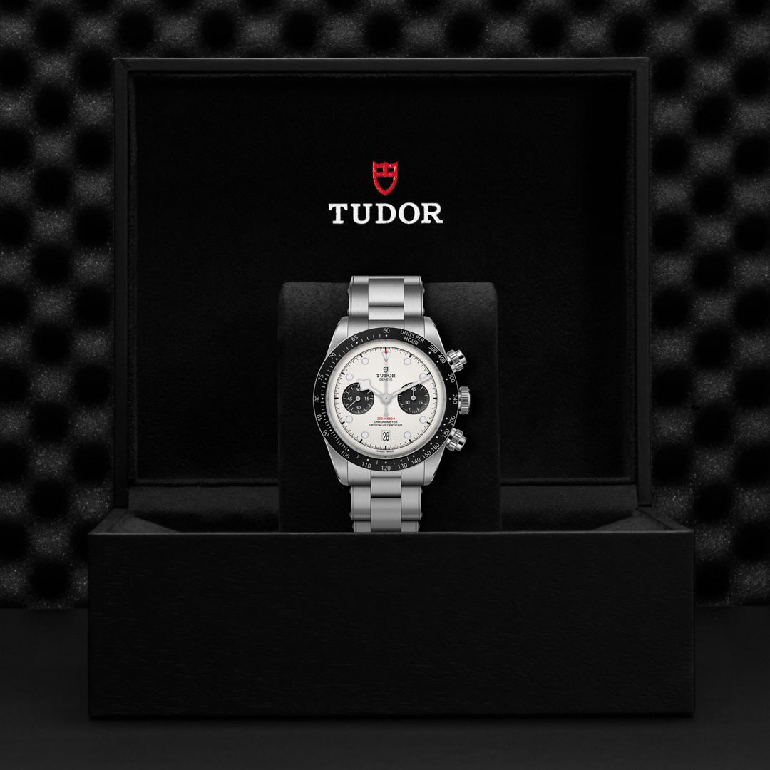 The TUDOR Black Bay Chrono, a luxury wristwatch with a silver band and white face featuring three sub-dials, is elegantly presented in an open black box. The inner lid displays the "TUDOR" brand name in red and white against the textured black background.