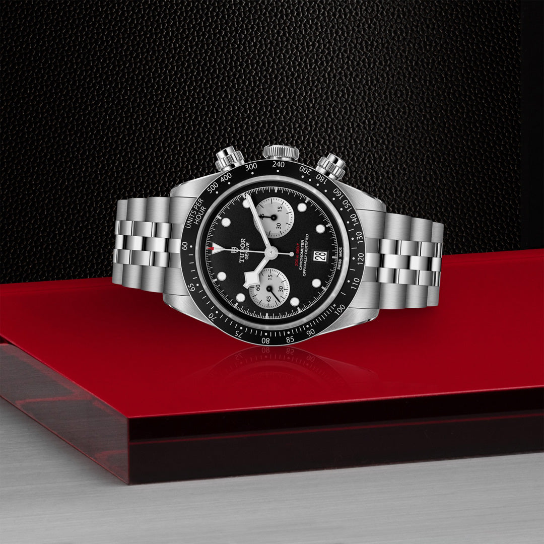 The TUDOR Black Bay Chrono, a luxury sports chronograph with a striking black dial and two white sub-dials, elegantly sits on a red and gray surface, featuring a metal bracelet against a textured black backdrop.