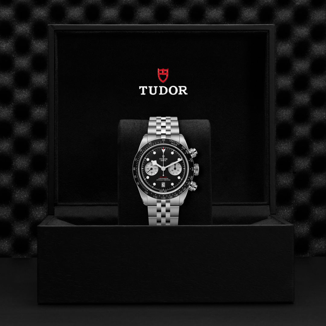 The TUDOR Black Bay Chrono luxury watch features a silver chain band, black face with white dials, and silver markers. It's secured by a T-Fit clasp and presented in a black box with the TUDOR logo on the lid.