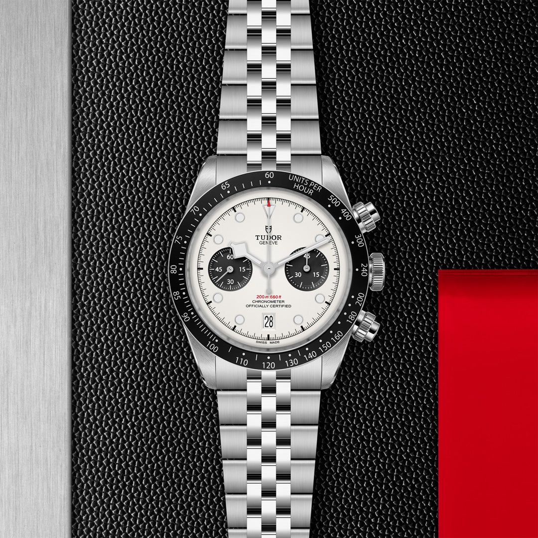 The TUDOR Black Bay Chrono is a luxury wristwatch featuring a silver metal bracelet, round white face, black sub-dials, and a black bezel with white numerals. It includes a T-Fit clasp and is elegantly set on a textured black and red surface.