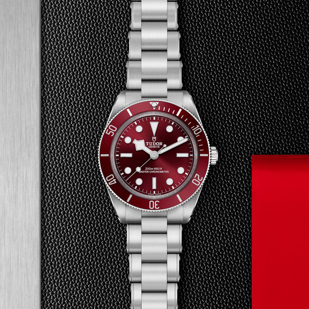 The TUDOR Black Bay 58 wristwatch showcases a maroon bezel and black dial with white markers. It features a silver metal bracelet, set against a textured black and red background, displaying the upright brand logo and details.