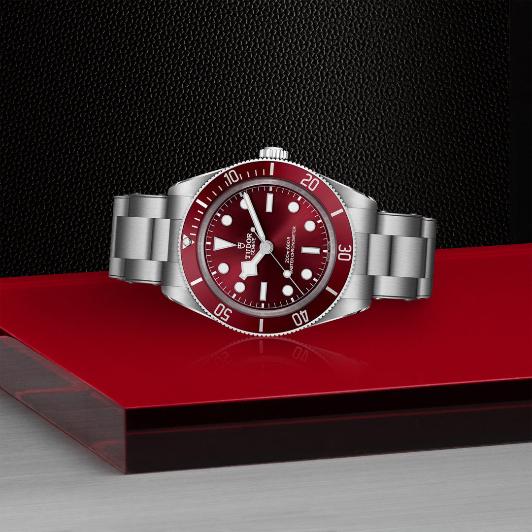A TUDOR Black Bay 58 dive watch, showcasing a burgundy dial and bezel, rests on a red and silver surface. With white hour markers, luminous hands, and a date window set against a textured black backdrop, it exemplifies style and precision.