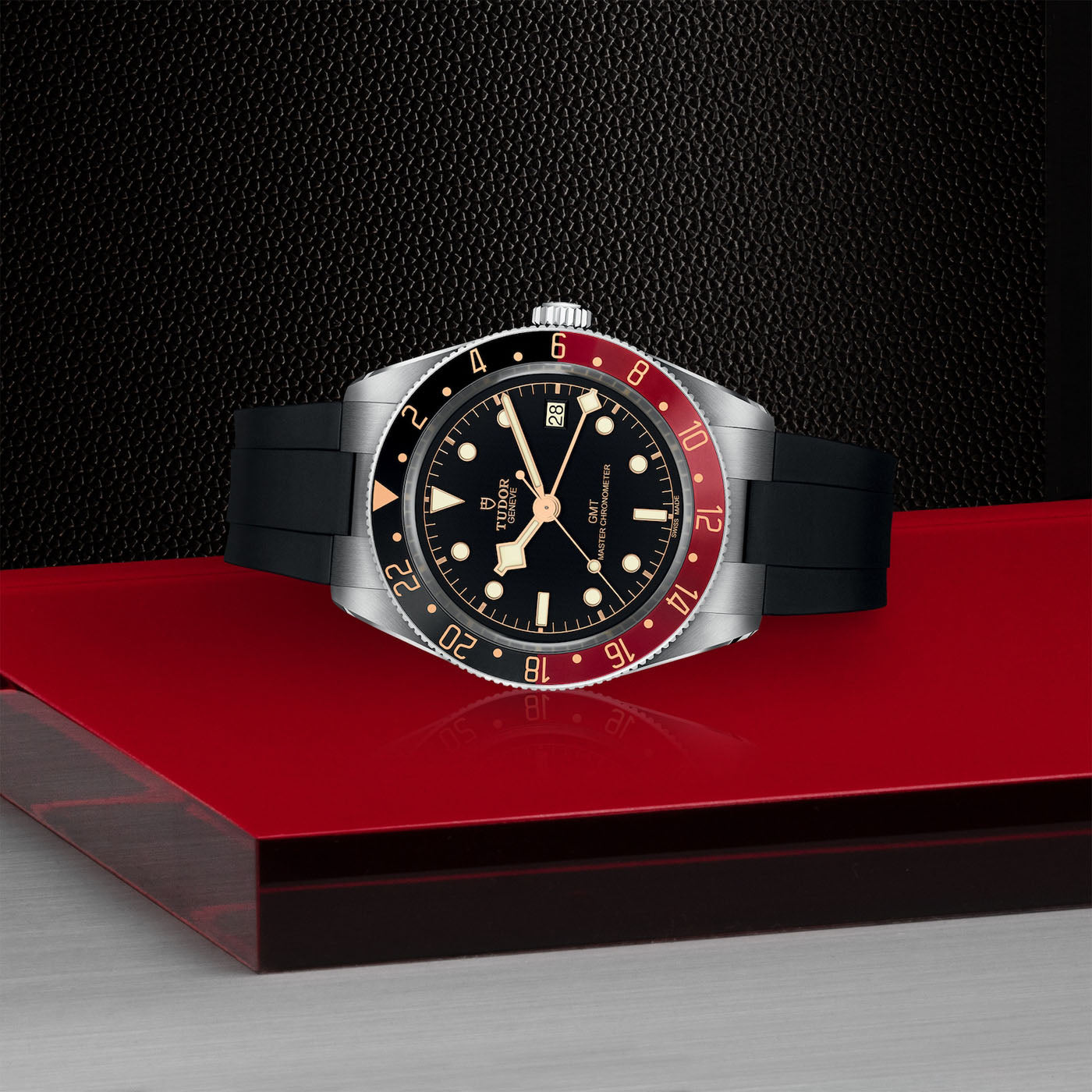 A luxury TUDOR Black Bay 58 GMT dive watch, featuring a black dial, red and black bezel, and a black strap, is displayed on a glossy red surface against a textured dark background. The watch shows the time as 10:10 and the date as 25.