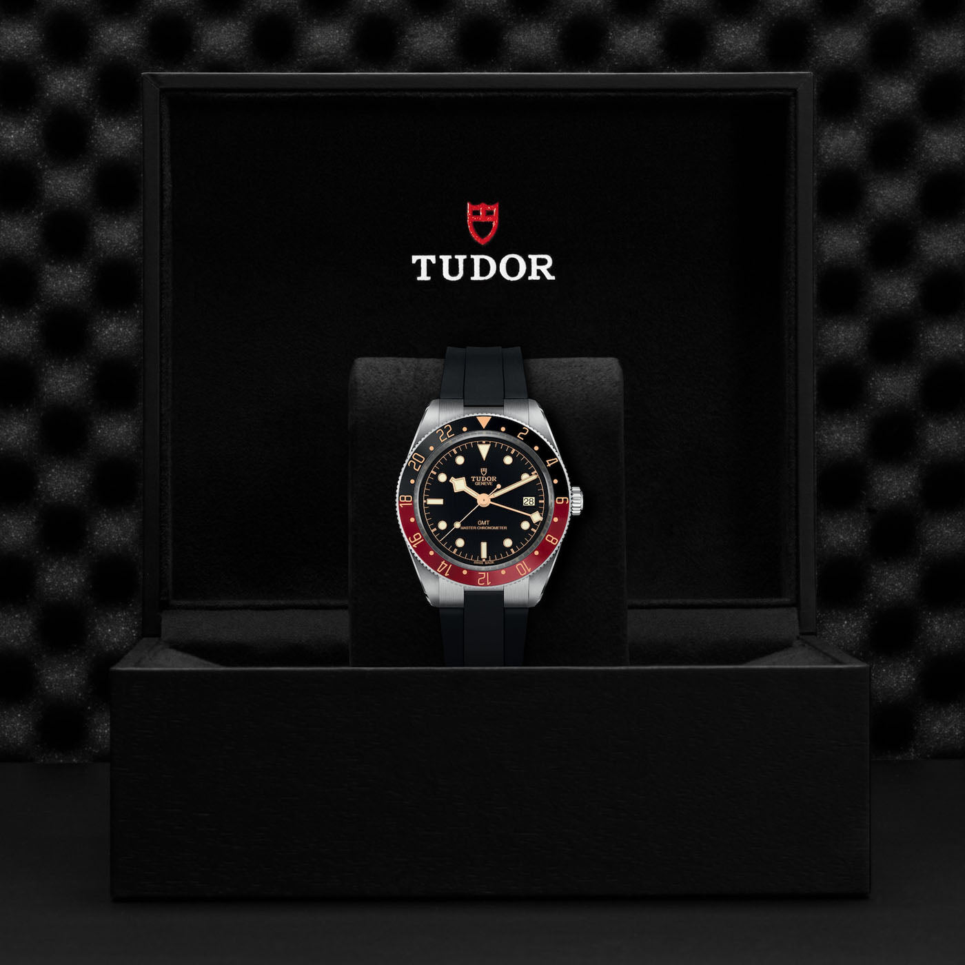 A TUDOR Black Bay 58 GMT, featuring a black and red bezel, is elegantly showcased in an open black box with a velvet interior. The prominently displayed TUDOR dive watch logo above the timepiece emphasizes its sophistication.