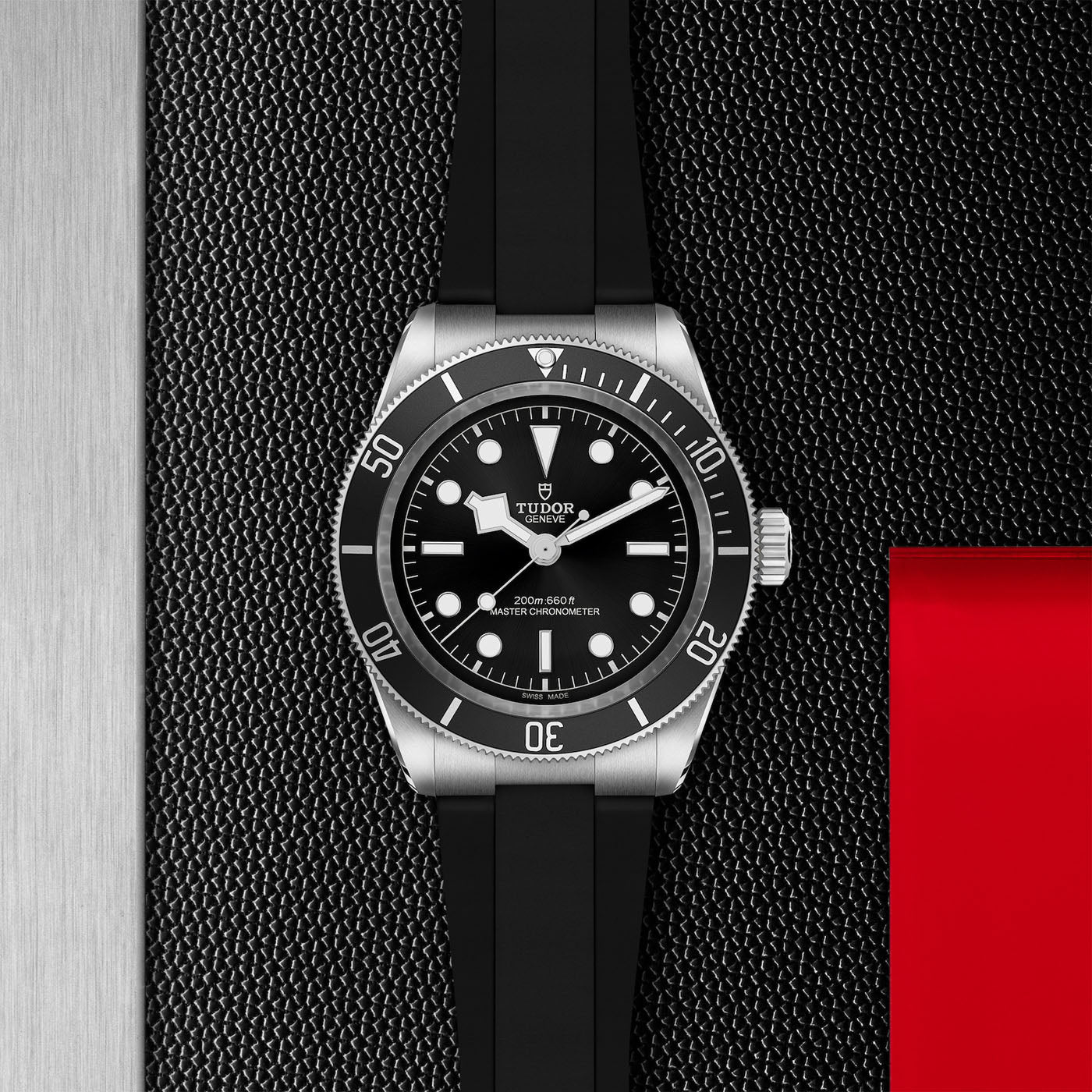 The TUDOR Black Bay wristwatch combines stylish neo-vintage design with a black dial and bezel, featuring white hour markers. It is complemented by a Black Bay-inspired black rubber strap and includes a red accent reminiscent of TUDOR divers’ watches, all showcased on a textured surface.