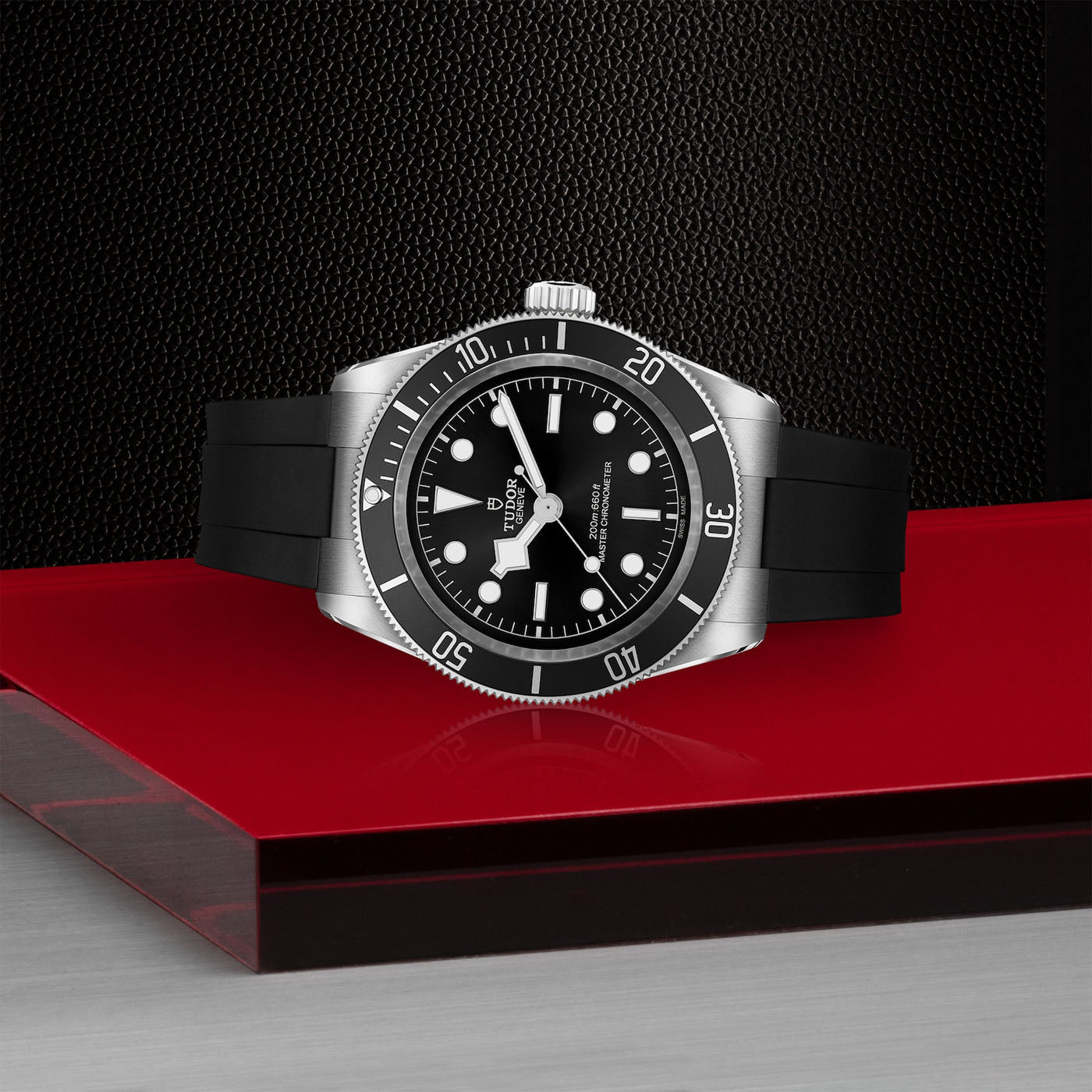 The TUDOR Black Bay, a neo-vintage luxury timepiece, showcases a black dial with silver accents as it rests elegantly on a glossy red surface against a textured backdrop. Drawing inspiration from TUDOR divers' watches, it boasts a black rubber strap and a stainless steel bezel adorned with numbers.