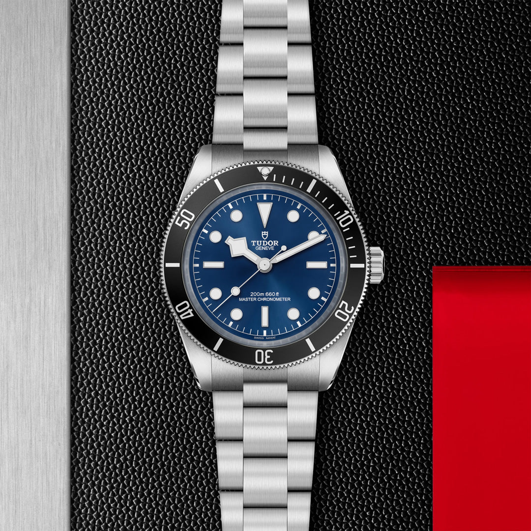 A TUDOR Black Bay 68 watch with a stainless steel case features a blue face, silver bezel, luminescent hour markers, and Snowflake hands, set against a textured black and metallic background. A red rectangle is partially visible on the right.