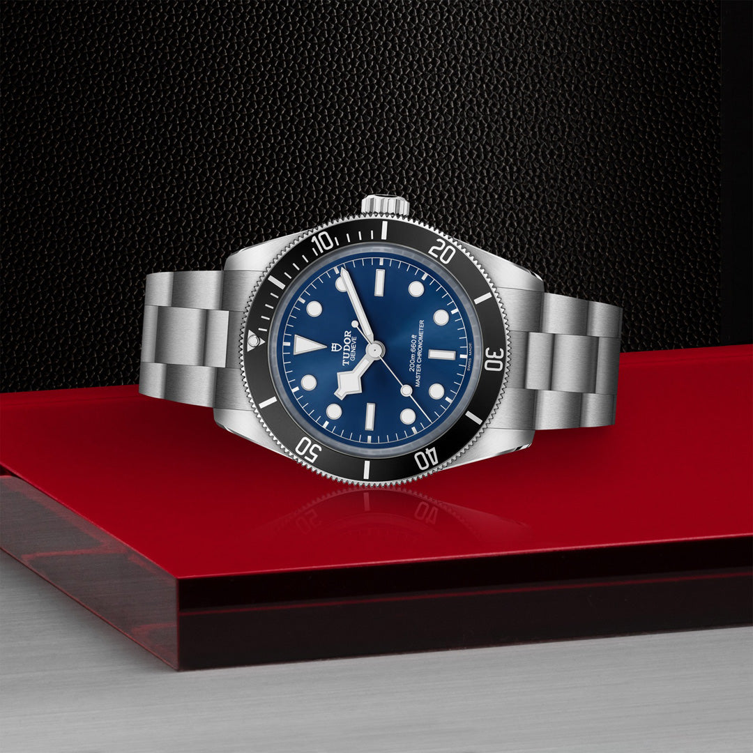 The TUDOR Black Bay 68, a stainless steel watch with snowflake hands, elegantly showcases a blue dial and black bezel on a red and gray surface against a textured black background. This Master Chronometer includes luminescent hour markers and a metal link bracelet.