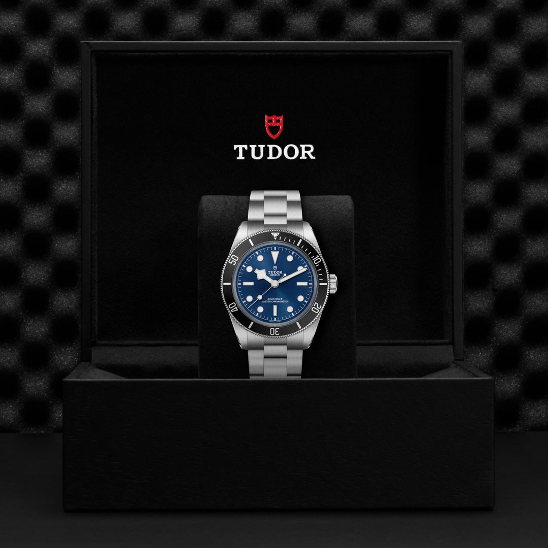 A TUDOR Black Bay 68 watch with a silver case, Snowflake hands, and a blue dial and bezel is elegantly presented in an open black box. The TUDOR logo adorns the inner lid above the watch, which rests on a textured black backdrop, exuding timeless sophistication.