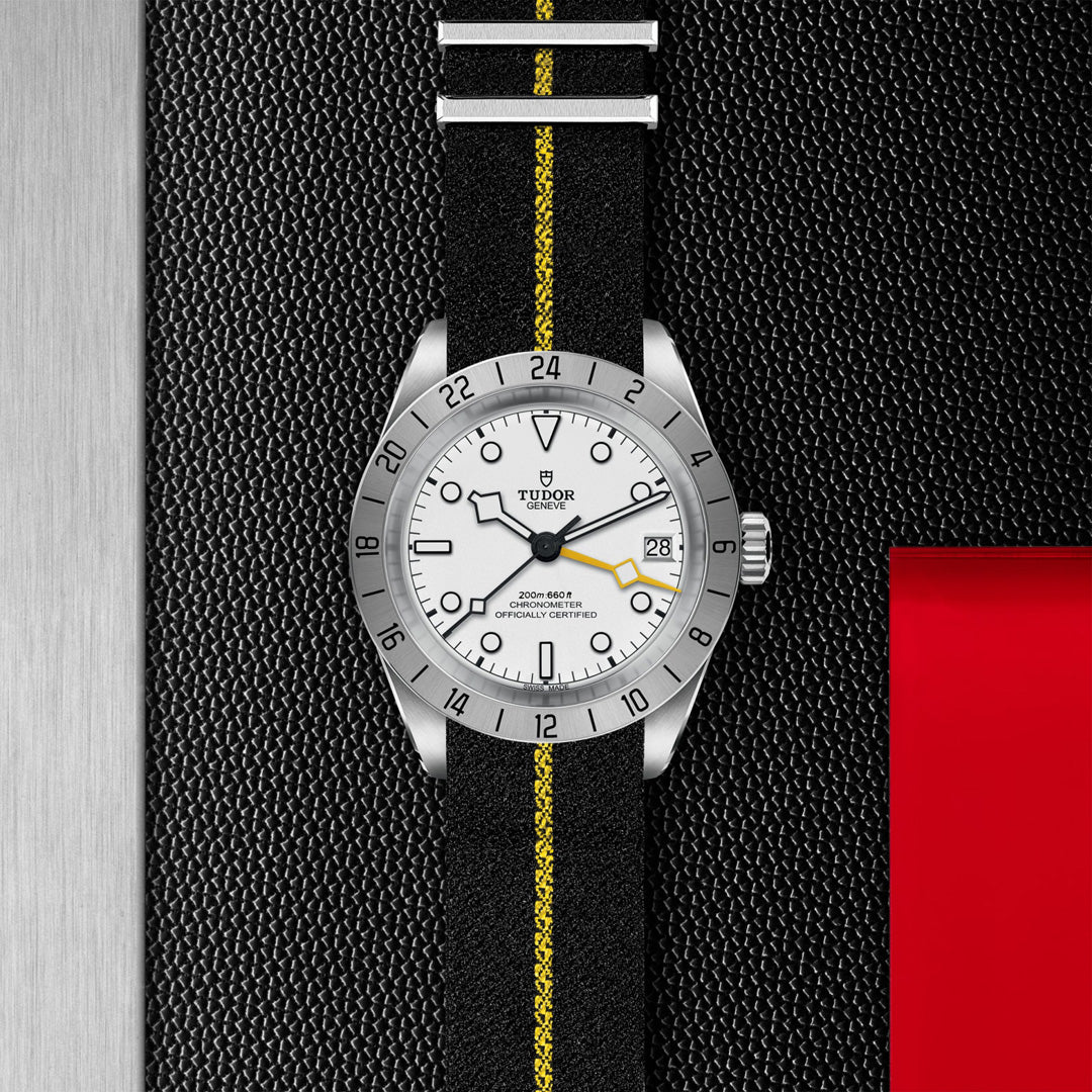 The TUDOR Black Bay Pro wristwatch features a silver bezel, white face with black hour markers and date display, powered by the TUDOR Manufacture Calibre MT5652. It includes a dual time zone function and is paired with a black fabric strap accented by a yellow stripe on a textured black panel with red highlights.