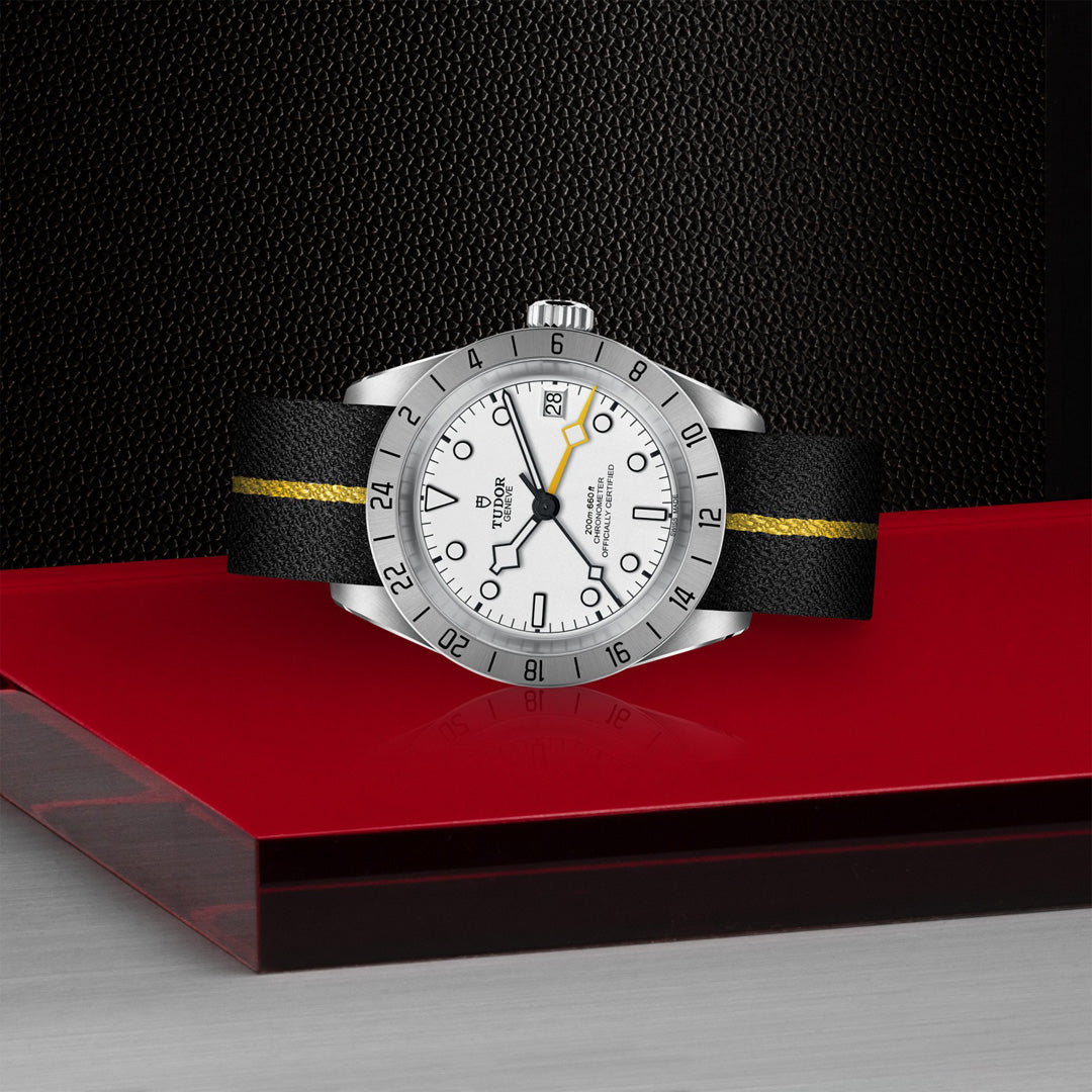 The TUDOR Black Bay Pro features a silver case with a white face and a black leather strap accented by a yellow stripe. It's equipped with dual time zone functionality, date display, and luminescent hour markers, resting on a red and black platform over a textured black background.