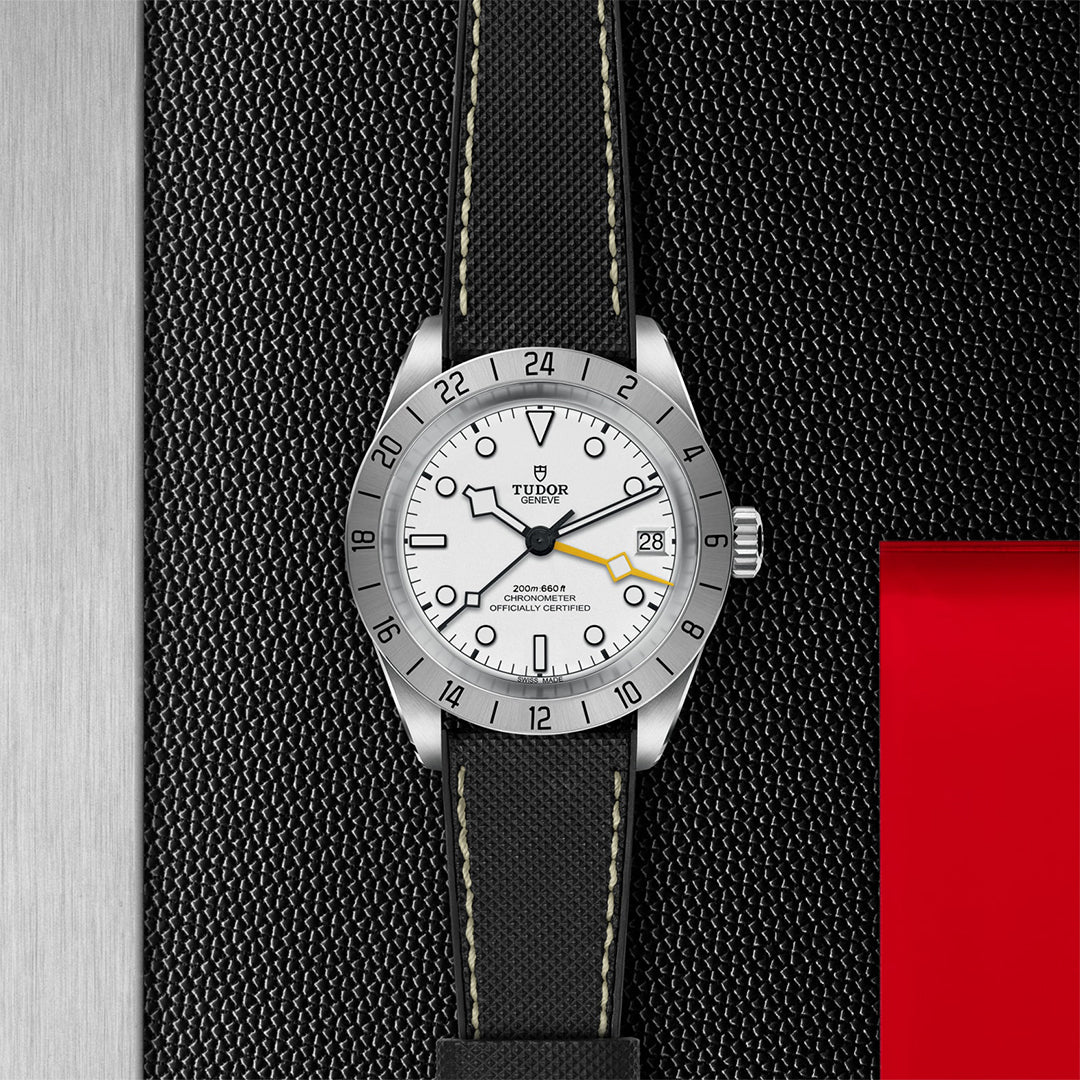 The TUDOR Black Bay Pro wristwatch features a silver bezel and white face with date display, complemented by a black leather strap with green stitching. Inspired by technical watches, the design sits on a textured black surface with silver and red accents.