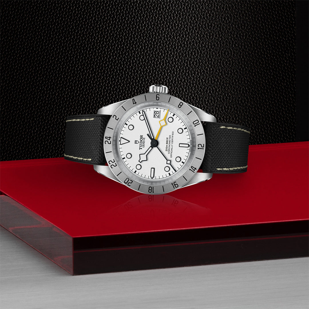 Set against a sleek black and red backdrop, the TUDOR Black Bay Pro features dual time zones with the TUDOR Manufacture Calibre MT5652. It has a stainless steel case, white face, black hour markers, a yellow arrow-tipped hand, and a 24-hour bezel with a date window at 3 o'clock.