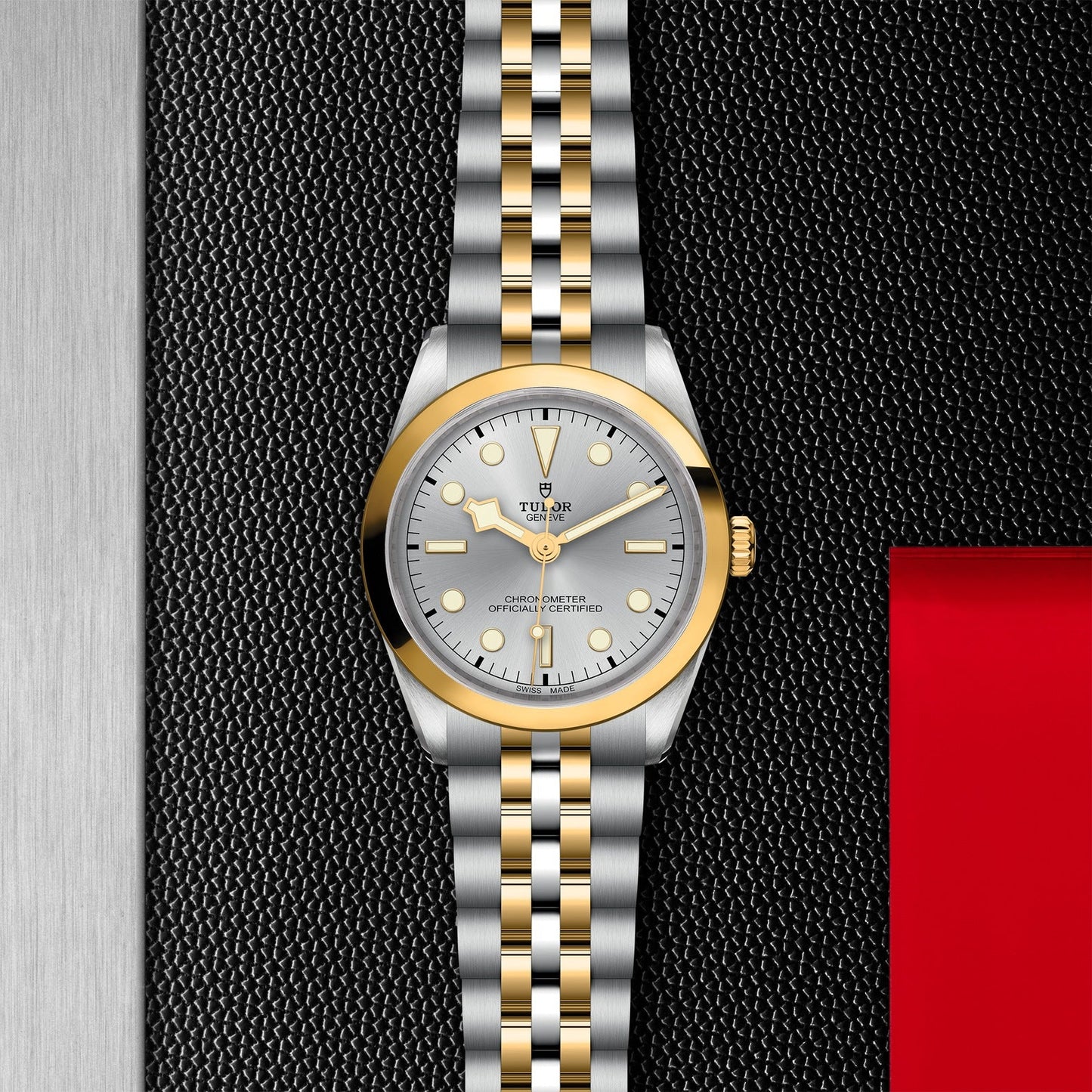 The TUDOR Black Bay 36 S&G wristwatch features a sophisticated silver and gold linked bracelet, enhanced by a round silver dial with gold accents and slender hour markers. This elegant timepiece is stylishly showcased on a textured black surface with understated red and silver background elements, drawing inspiration from the iconic Black Bay collection.