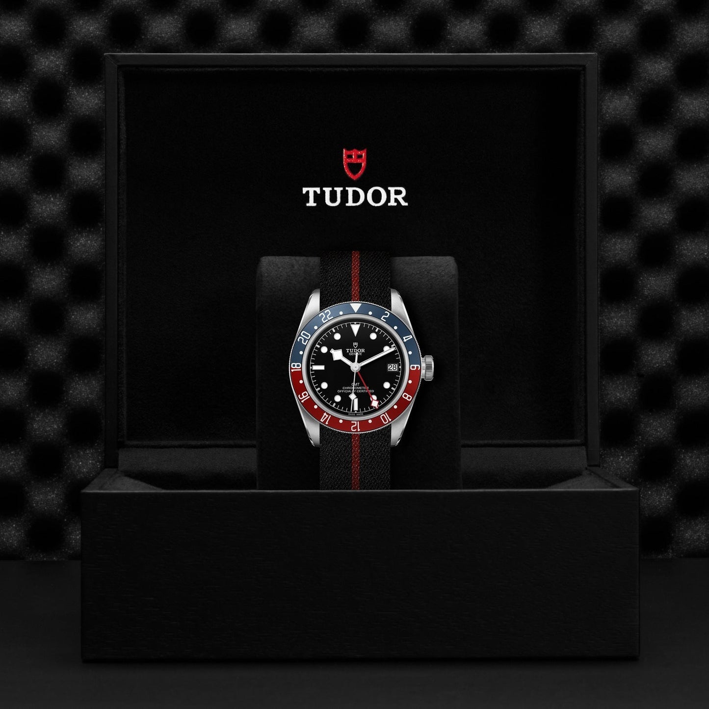 A TUDOR Black Bay GMT watch with a black fabric strap is showcased in an open black box. This timepiece boasts a rotatable blue and red bezel, accompanied by a black dial with white markers. The TUDOR logo is prominently displayed on the inner lid of the box, accentuating its timeless elegance and multiple time-zone functionality.