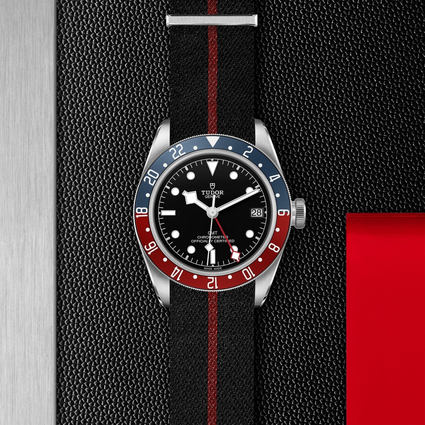 The TUDOR Black Bay GMT, a luxurious wristwatch, showcases a black dial accented by a bold red and blue rotatable bezel. It features a stylish black NATO strap with a central red stripe, elegantly set against a textured metallic design with red accents.